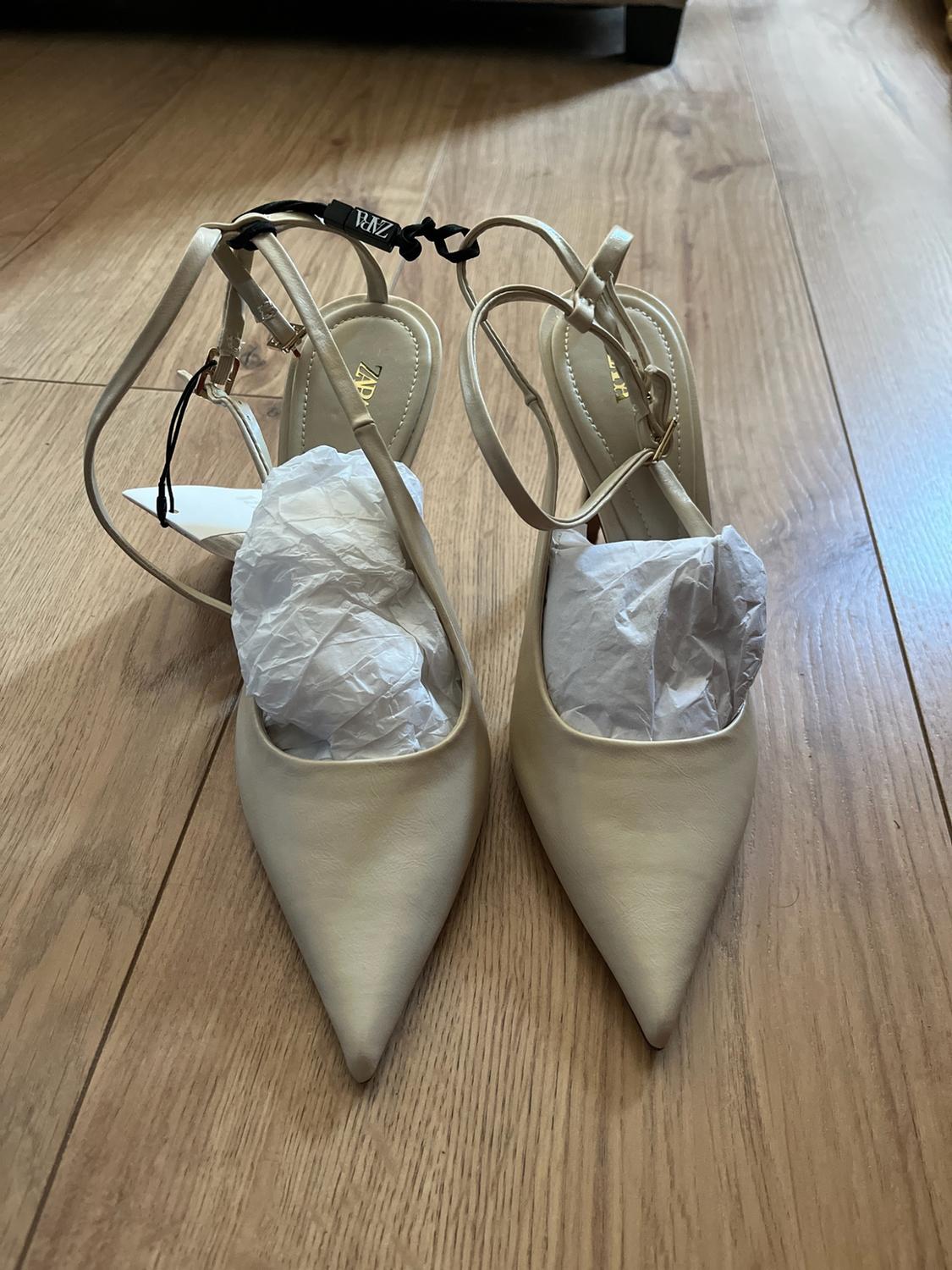 Beige pointed-toe heeled shoes with straps on a wooden floor. They have paper inside and a black label with the text ZARA.