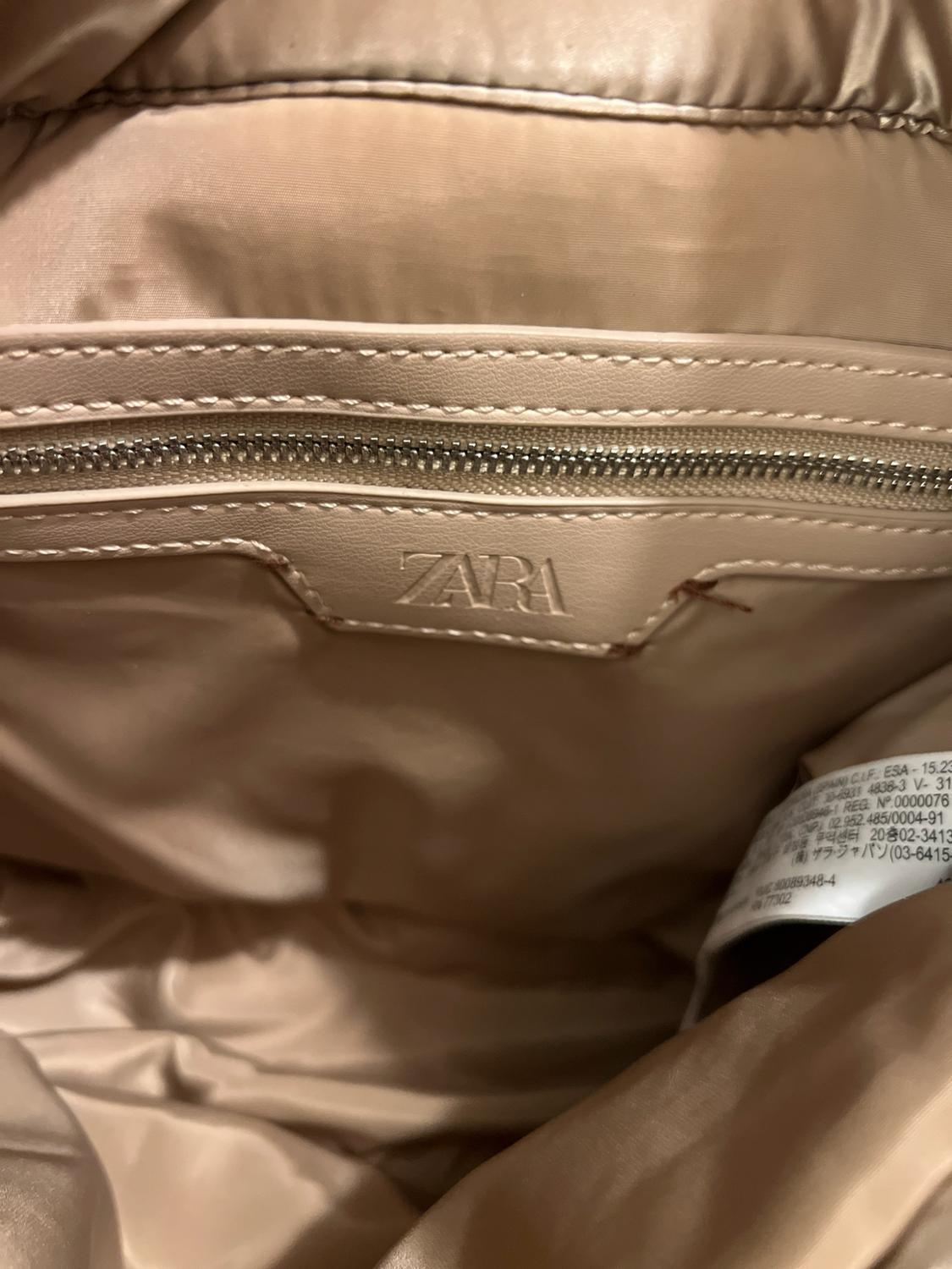 Detail of the inside of a beige bag. A zippered pocket, care labels, and a patch bearing the text ZARA can be seen.