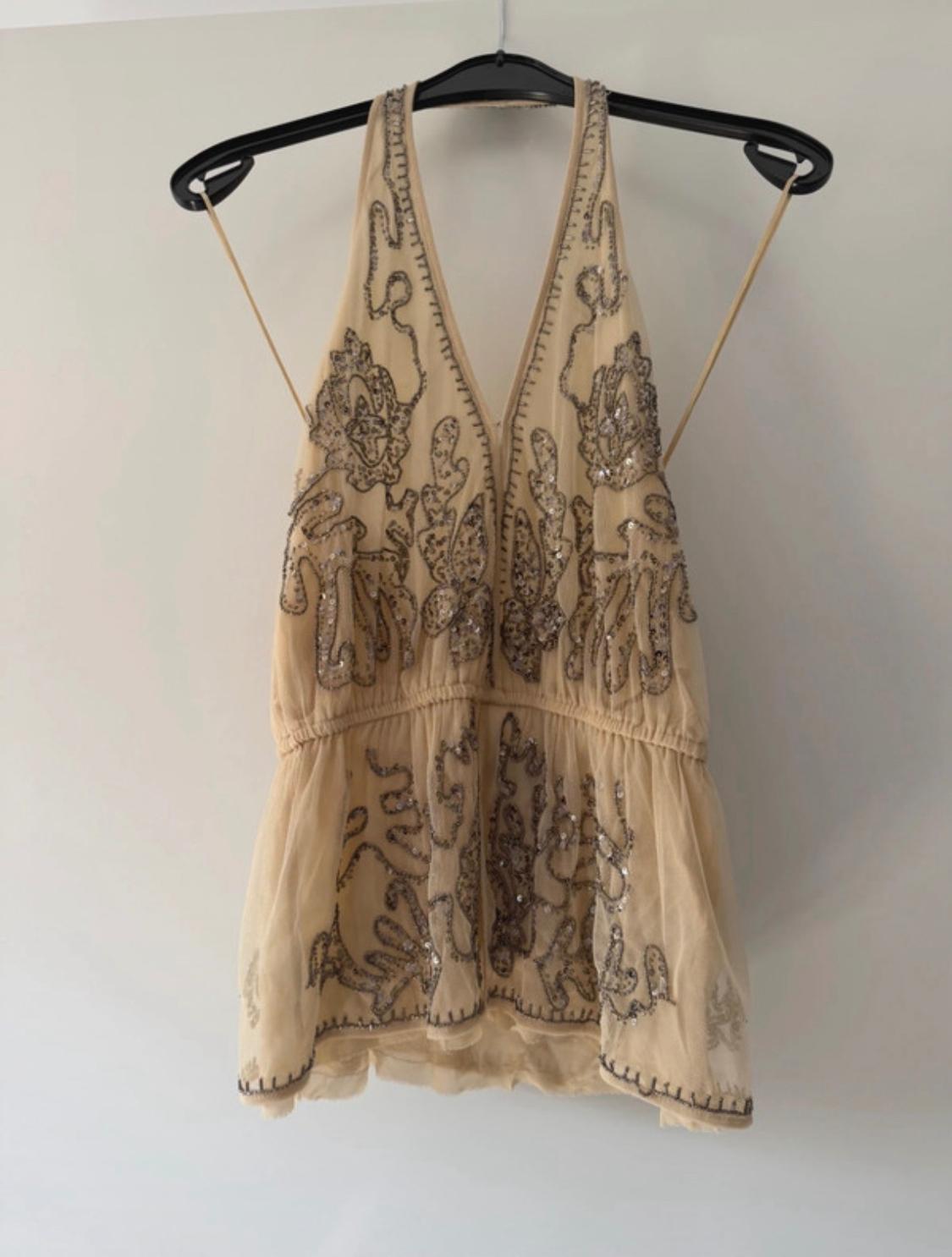 Beige top with a V-neckline, thin straps and silver sequin embroidery, hanging on a black hanger against a white background.
