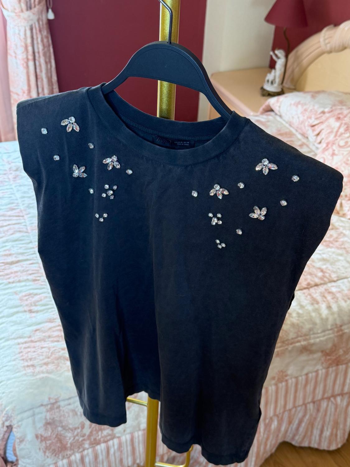Black T-shirt with sparkly floral embellishments, hanging on a gold hanger in a classically decorated bedroom.