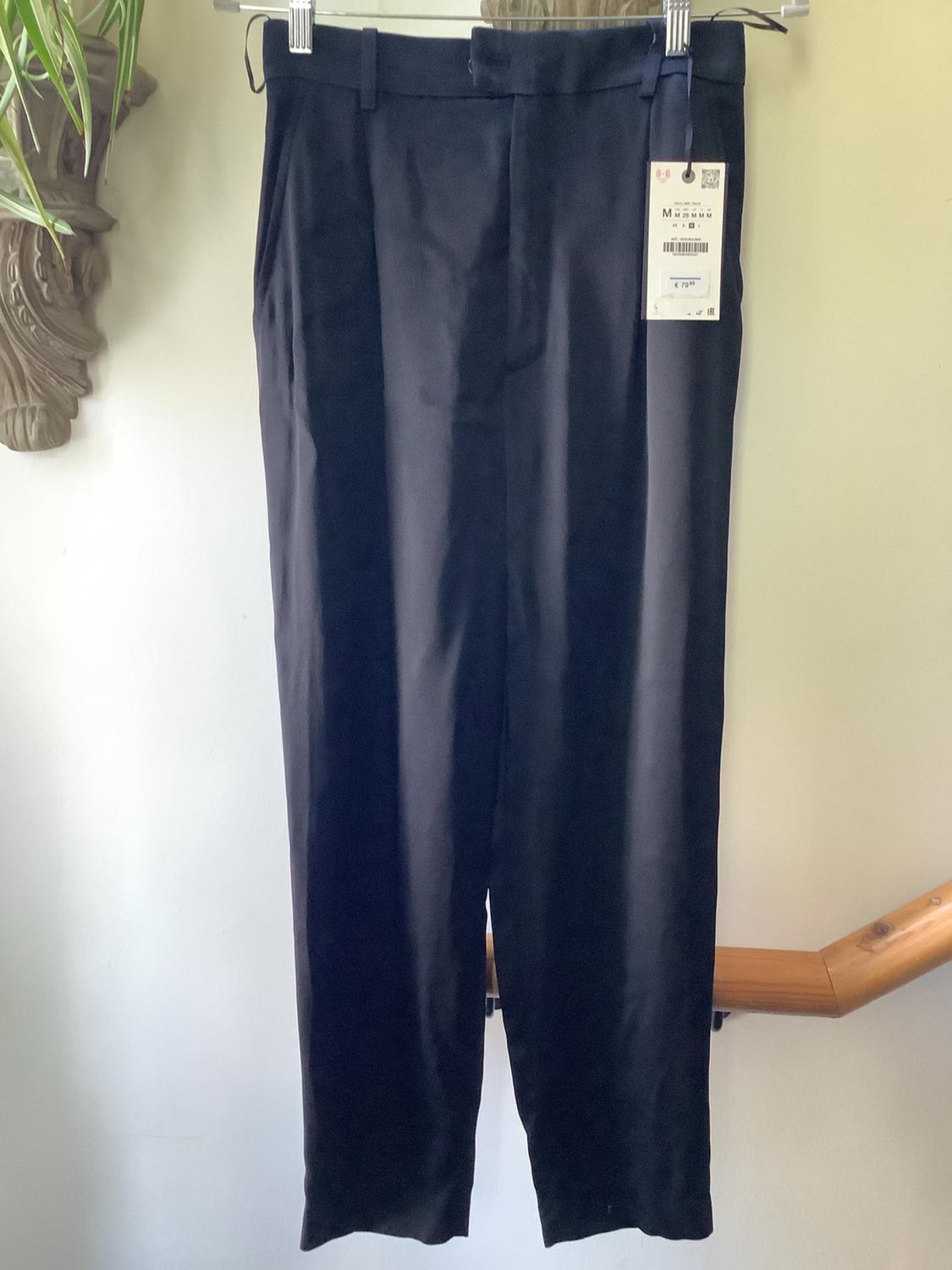 Black trousers hanging on a hanger with a visible tag. White background with wooden details and a decorative plant on the left.