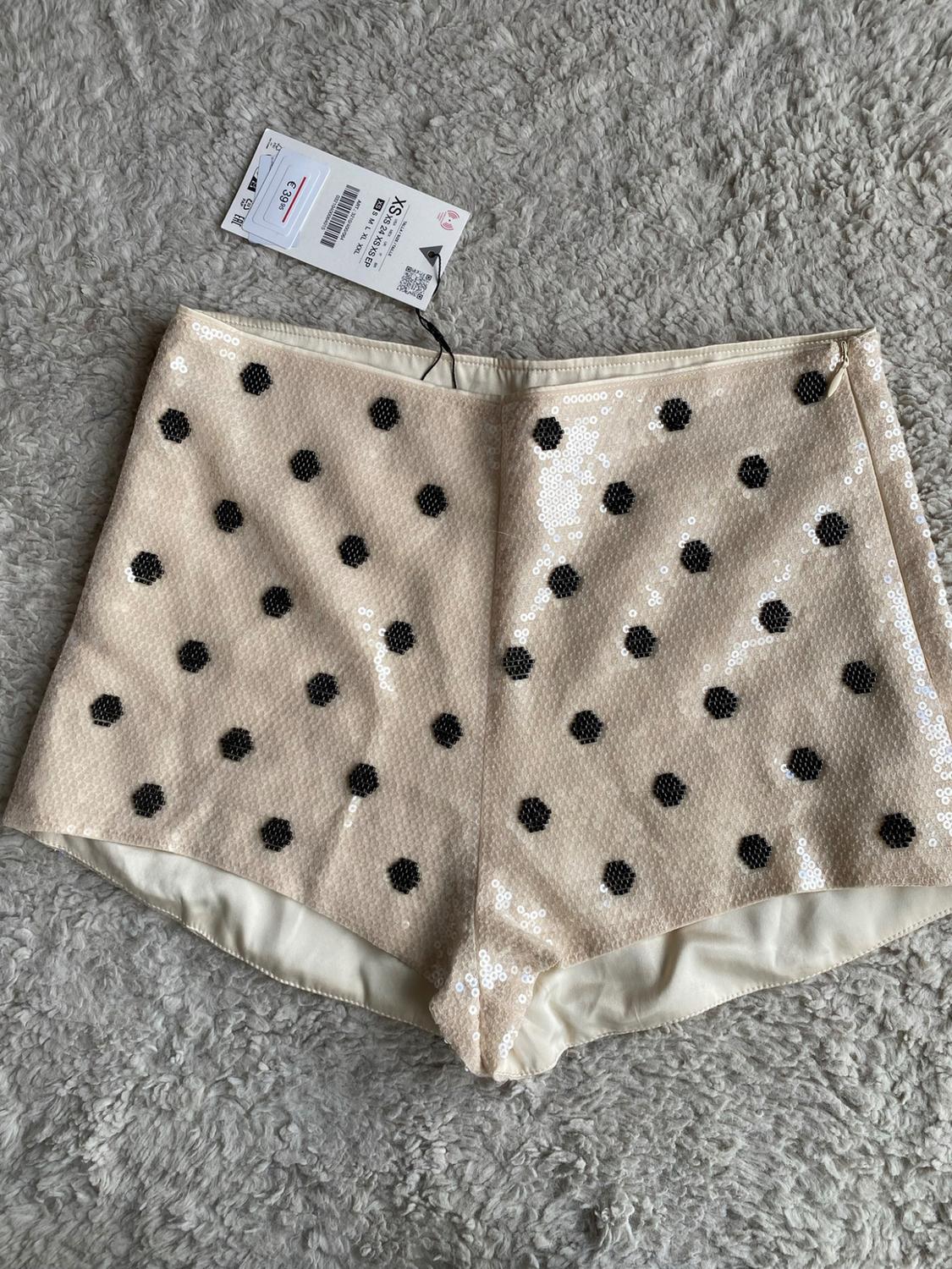 Beige sequin shorts with black polka dots on a shaggy rug. Labels with text: XS, €39.95.