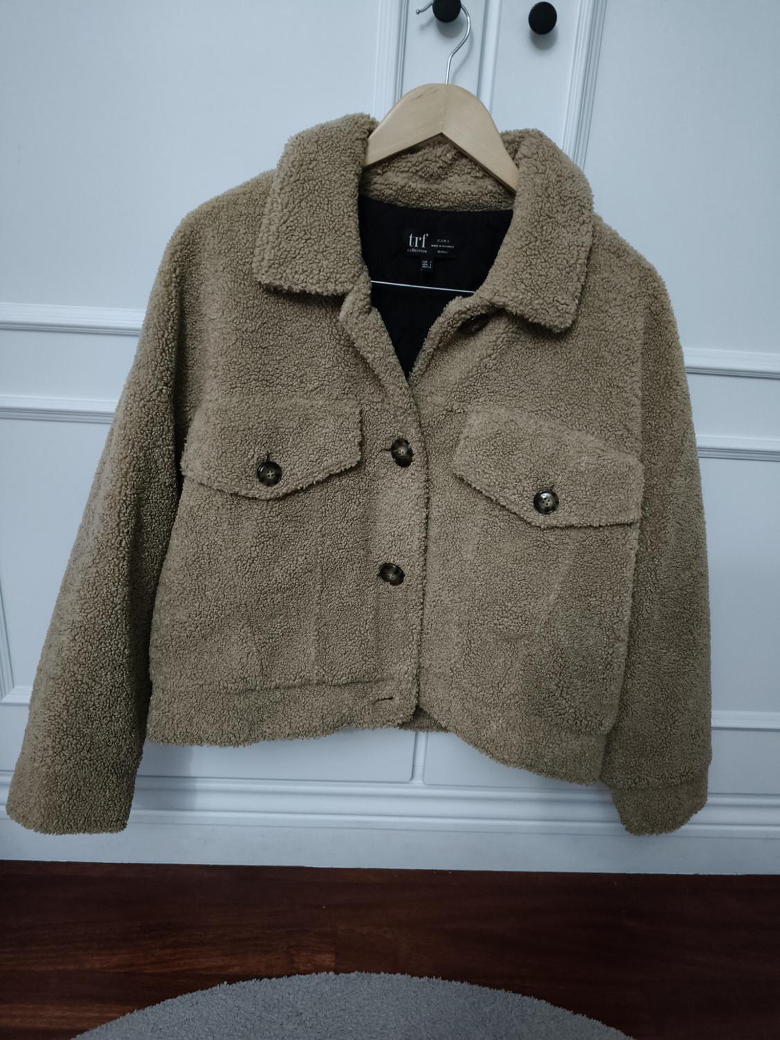 Beige teddy coat hung on a wooden hanger. It has two buttoned pockets on the front. White wall background.