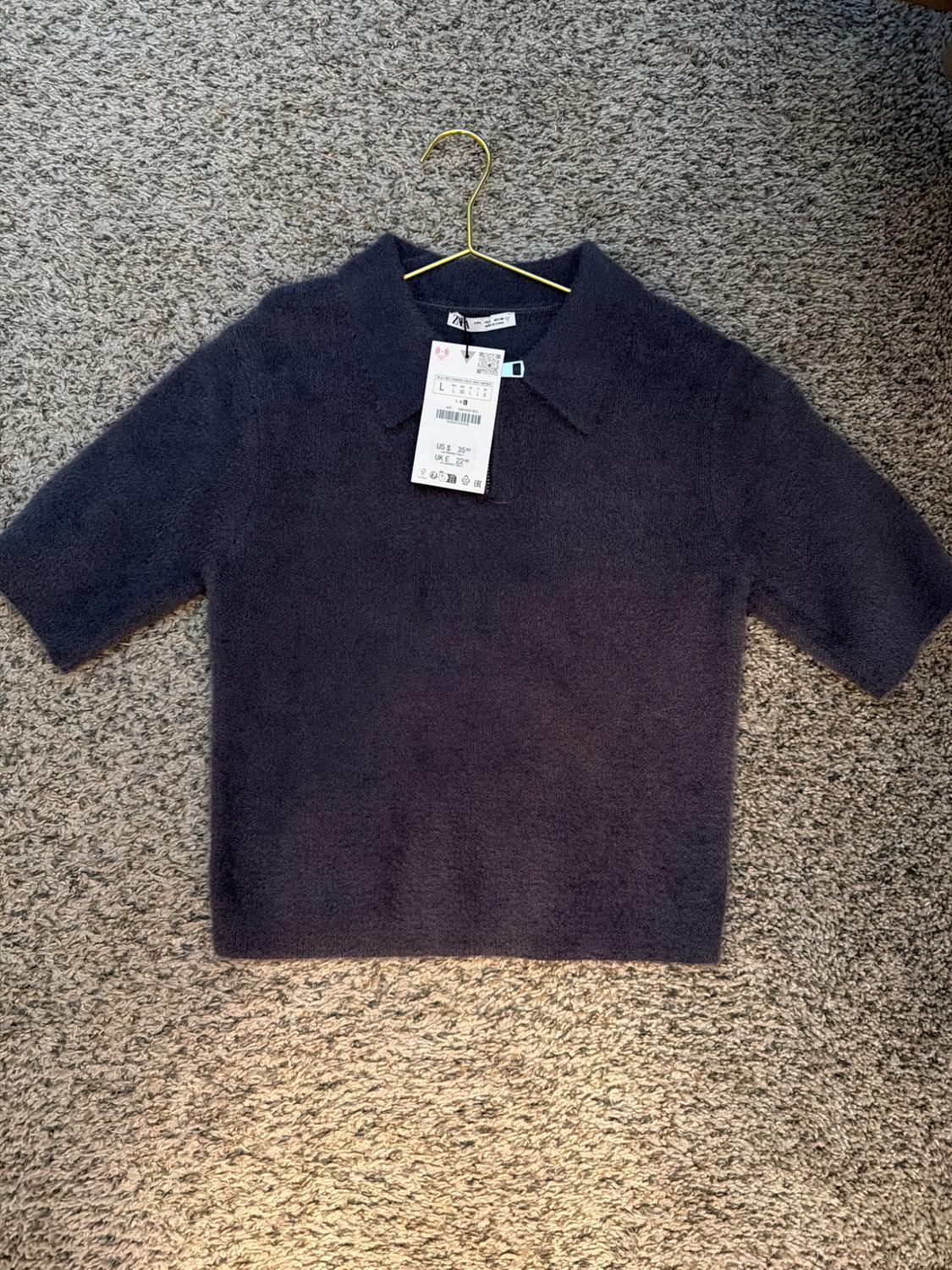 Dark blue short-sleeved collared jersey, hanging on a gold hanger over a gray carpet. Visible tag with size details.