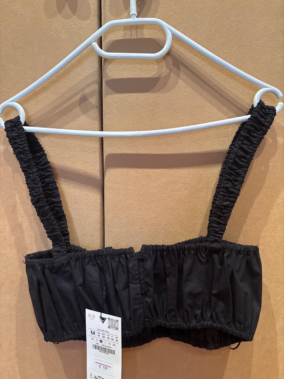 Black shirred-strap top hanging on a white hanger. Visible tag with a price of €19.99. Beige background.