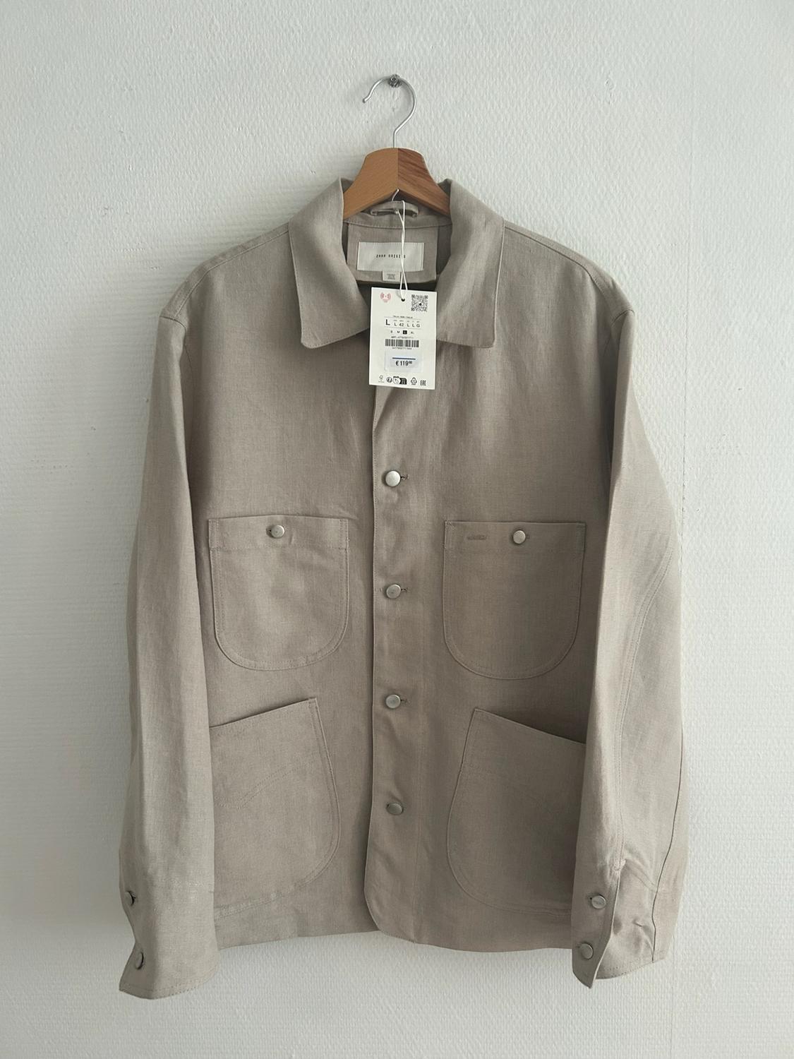 Beige overshirt with buttons and four front pockets, hanging on a wooden hanger against a white wall.