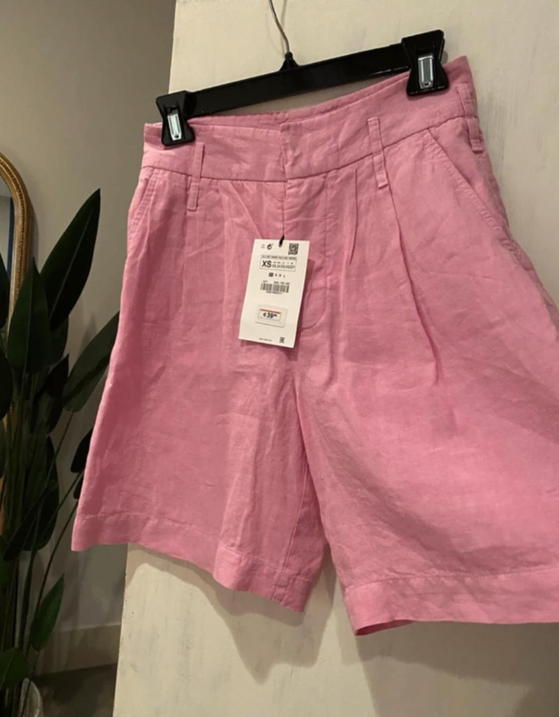 Pink shorts hanging on a black hanger with an XS size tag. Background with plant and partially visible mirror.