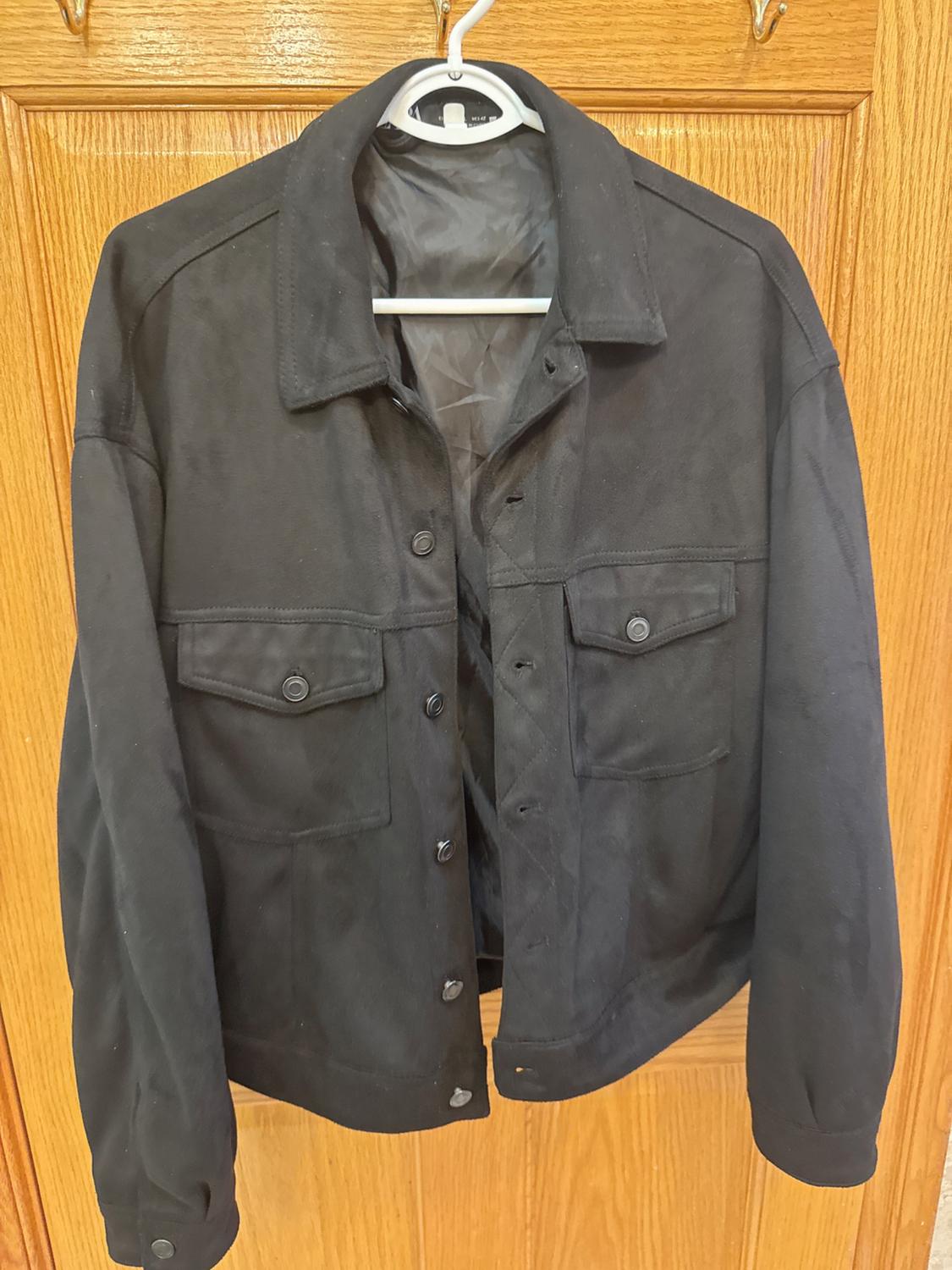 Black jacket hanging on a white hanger over a wooden door. It has flap pockets and front buttons.