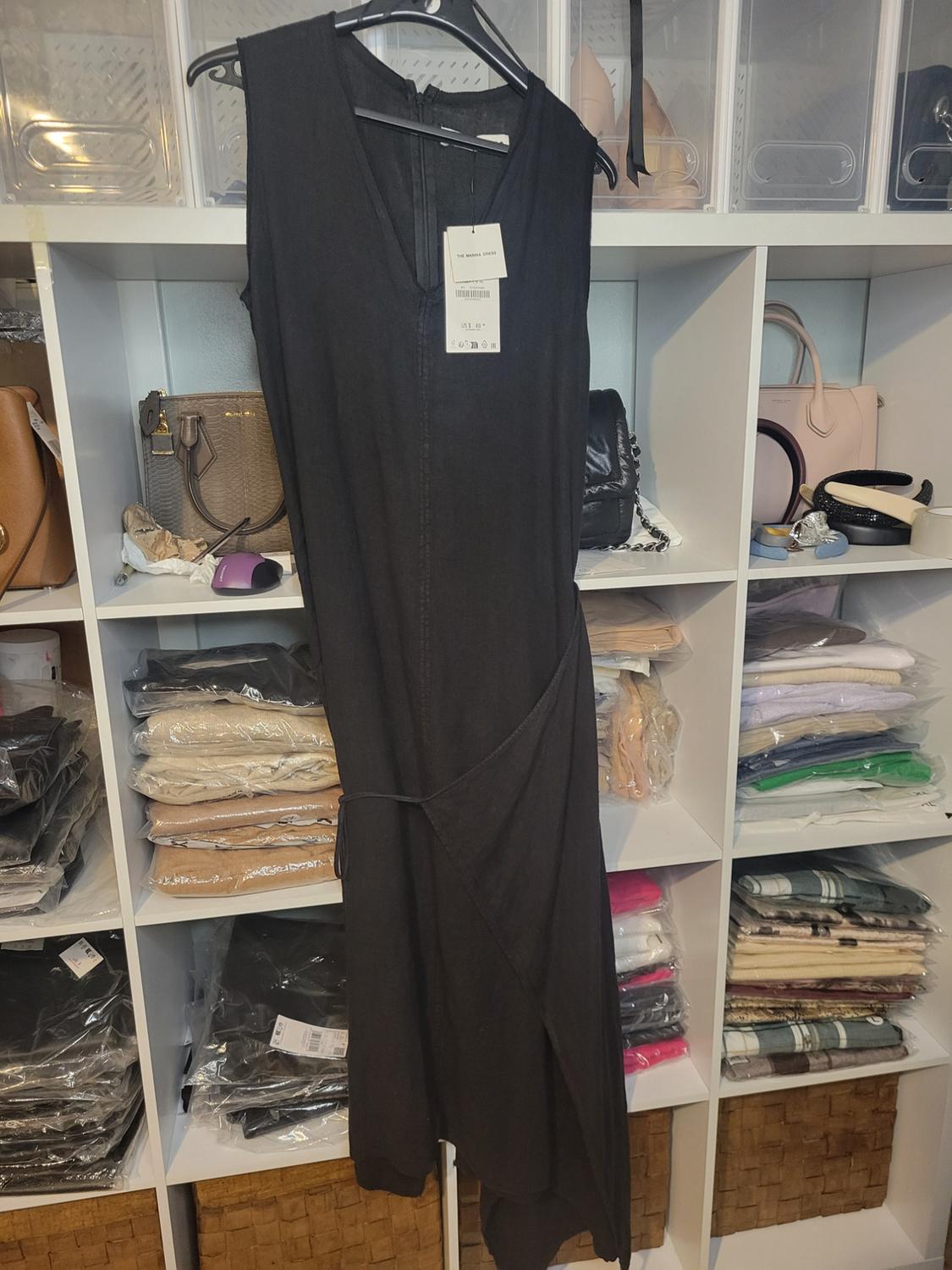 Sleeveless black dress hanging on a hanger, with visible tag, in front of a shelf with handbags and folded clothes.