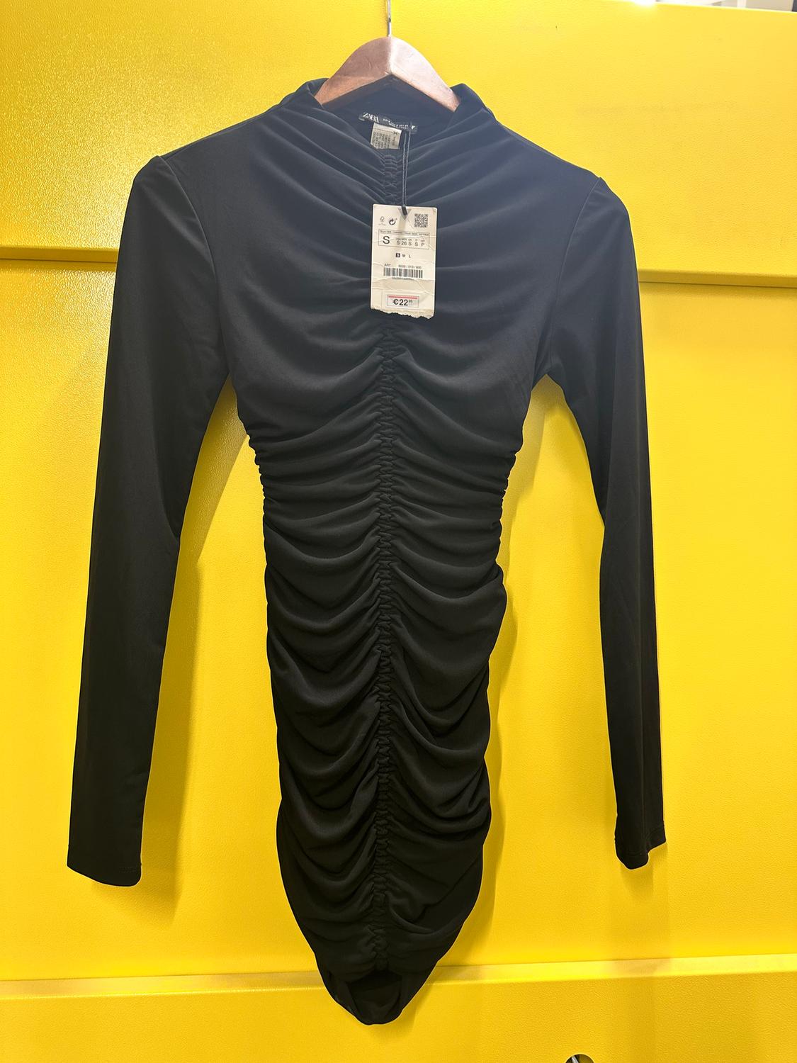 Black long-sleeved ruched dress hanging on a wooden hanger against a yellow background. Visible tag with price of €22.