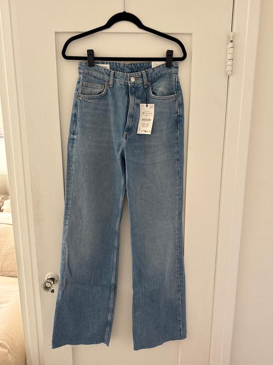 Wide-leg blue jeans hanging on a black hanger over a white door. Visible tag on the front pocket.
