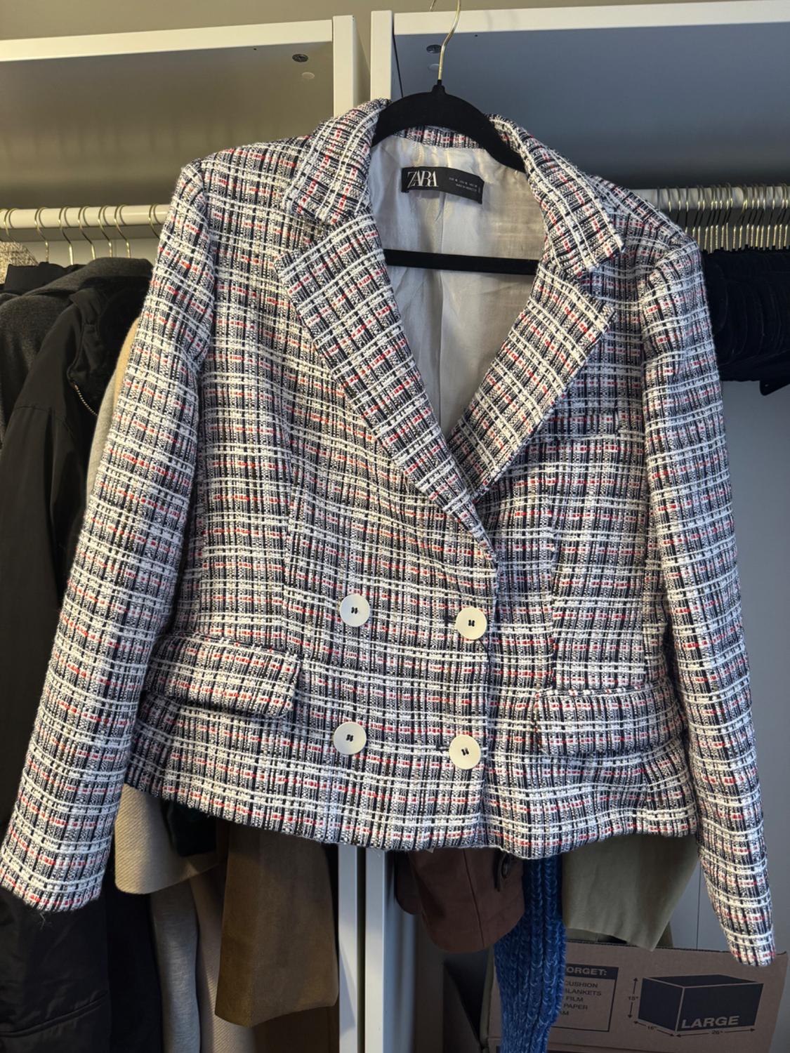 Checked jacket hanging in a wardrobe, with white buttons and a visible Zara label. Background with other hanging garments.