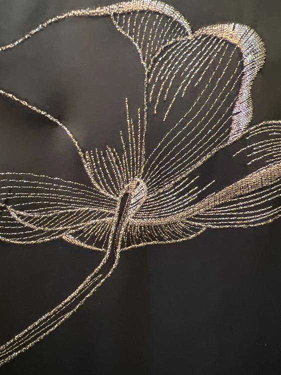 Floral design embroidered in gold thread on a black background, showing a detailed pattern of petals and a curved stem.