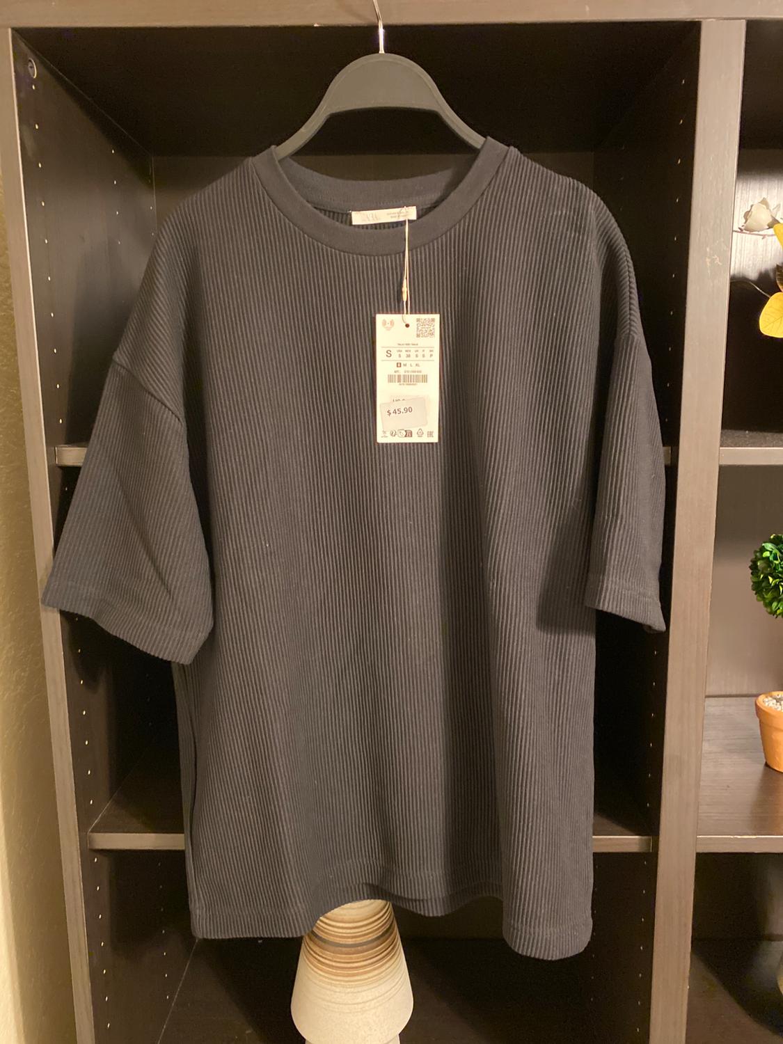 Black short-sleeved t-shirt with ribbed texture, hanging on a hanger inside a wardrobe. Visible tag with price of $45.90.