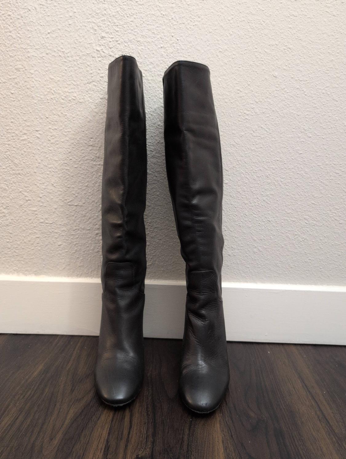 Pair of black leather high boots on a dark wood floor in front of a white wall, creating a minimalist composition.