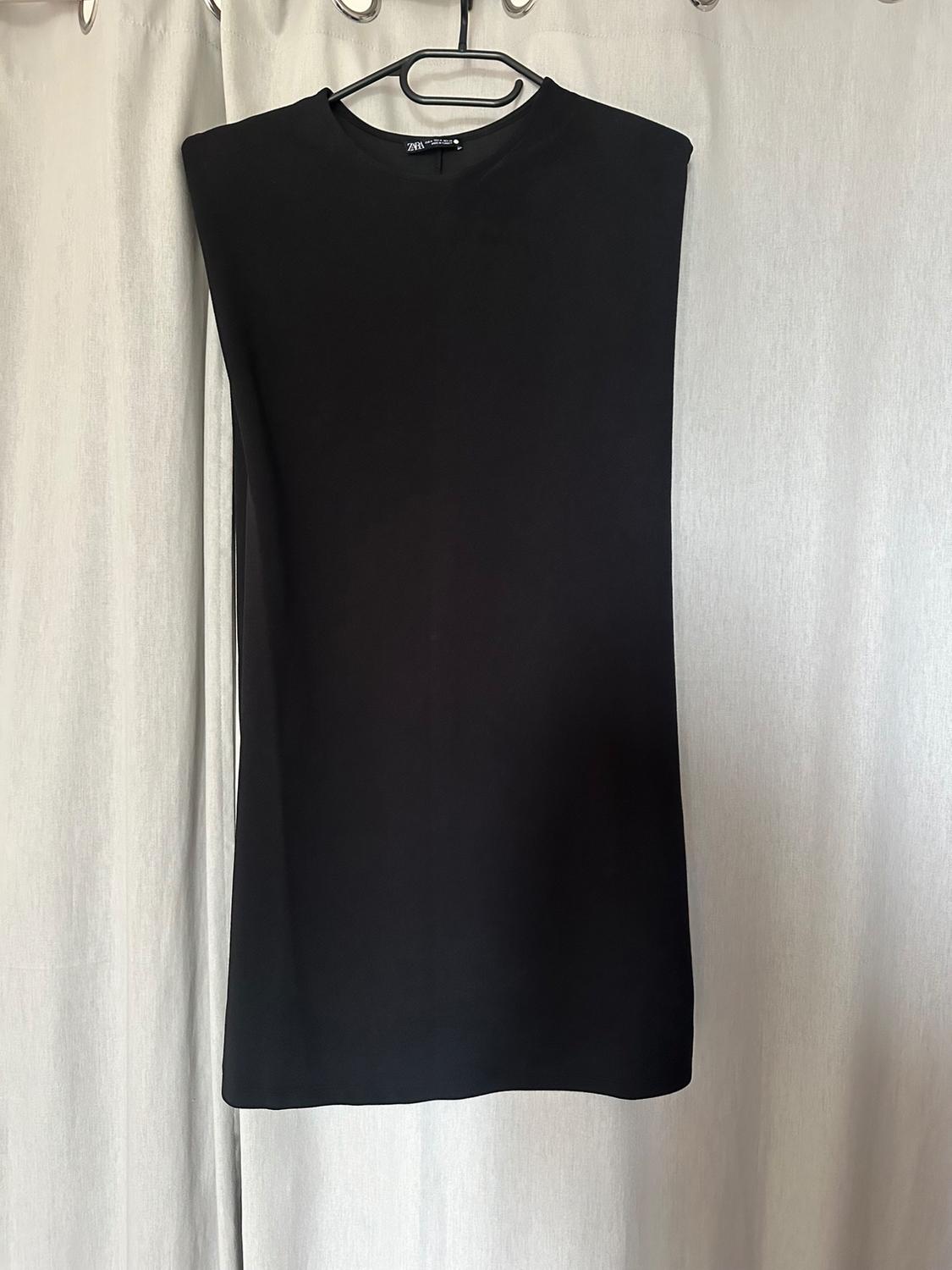 Sleeveless black dress hanging on a hanger in front of a white curtain. Label visible on the neck.