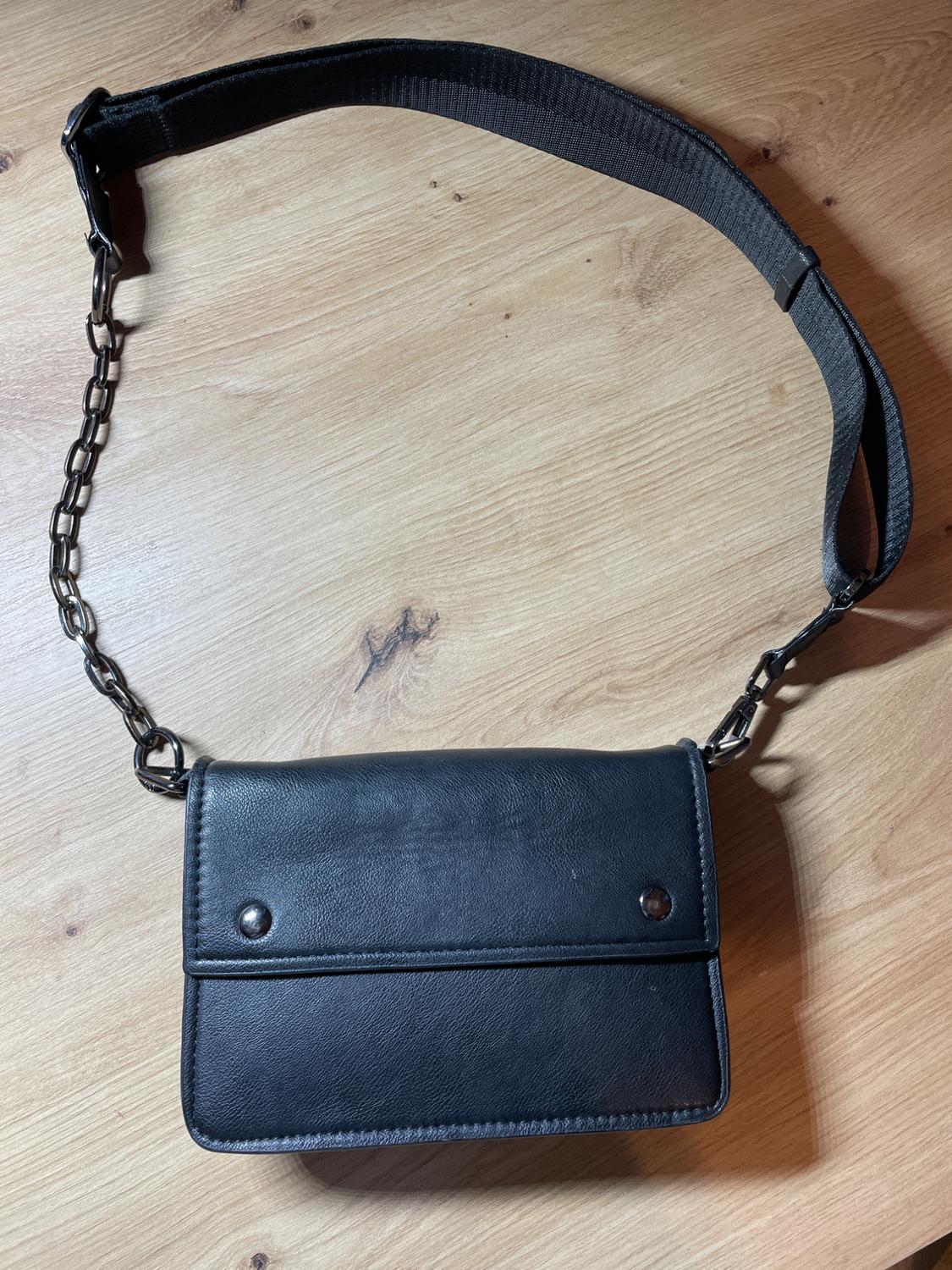 Black handbag with chain strap and fabric on a light wood background. Simple design with two studs on the flap.