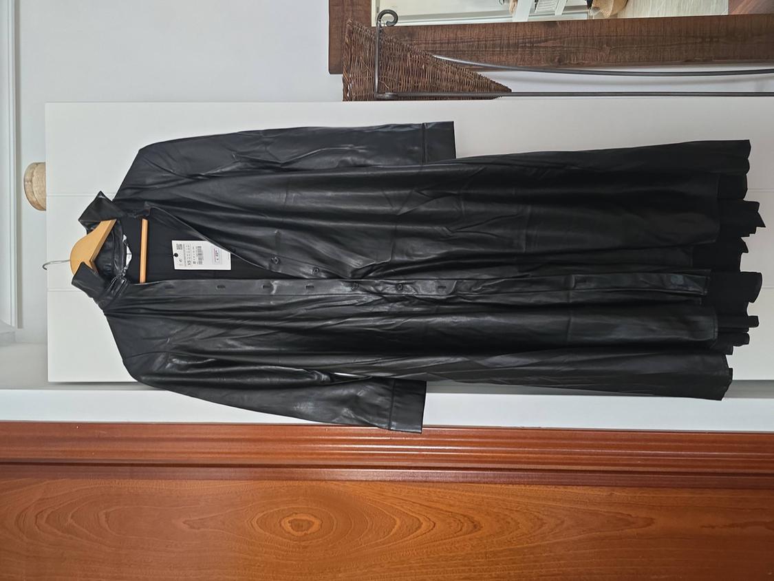 Black leather coat hanging on a wooden hanger over a wooden door. Label visible inside the coat.