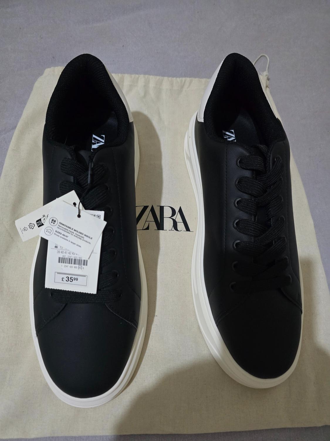 Black trainers with white soles on a bag. Label with text: ZARA, REMOVABLE MOLDED INSOLE, SIZES 46/47, £35.99.