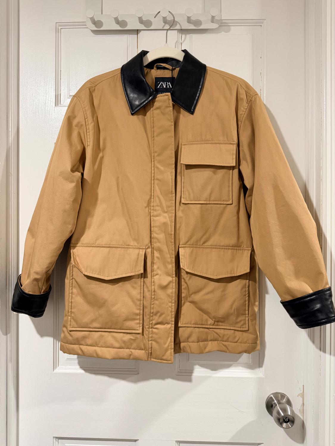 Beige jacket with black collar and cuffs hanging on a white door. Visible label with text "ZARA".