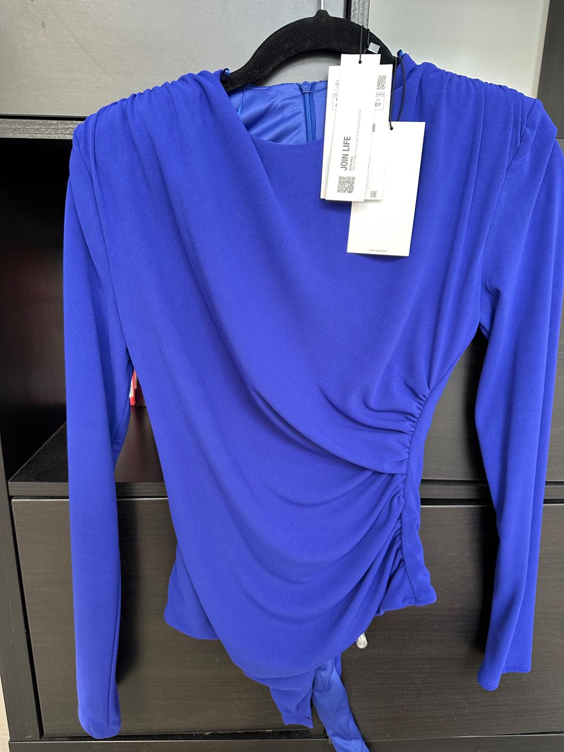 Blue blouse with draping and long sleeves hanging on a black hanger. White tags with "JOIN LIFE" text visible.