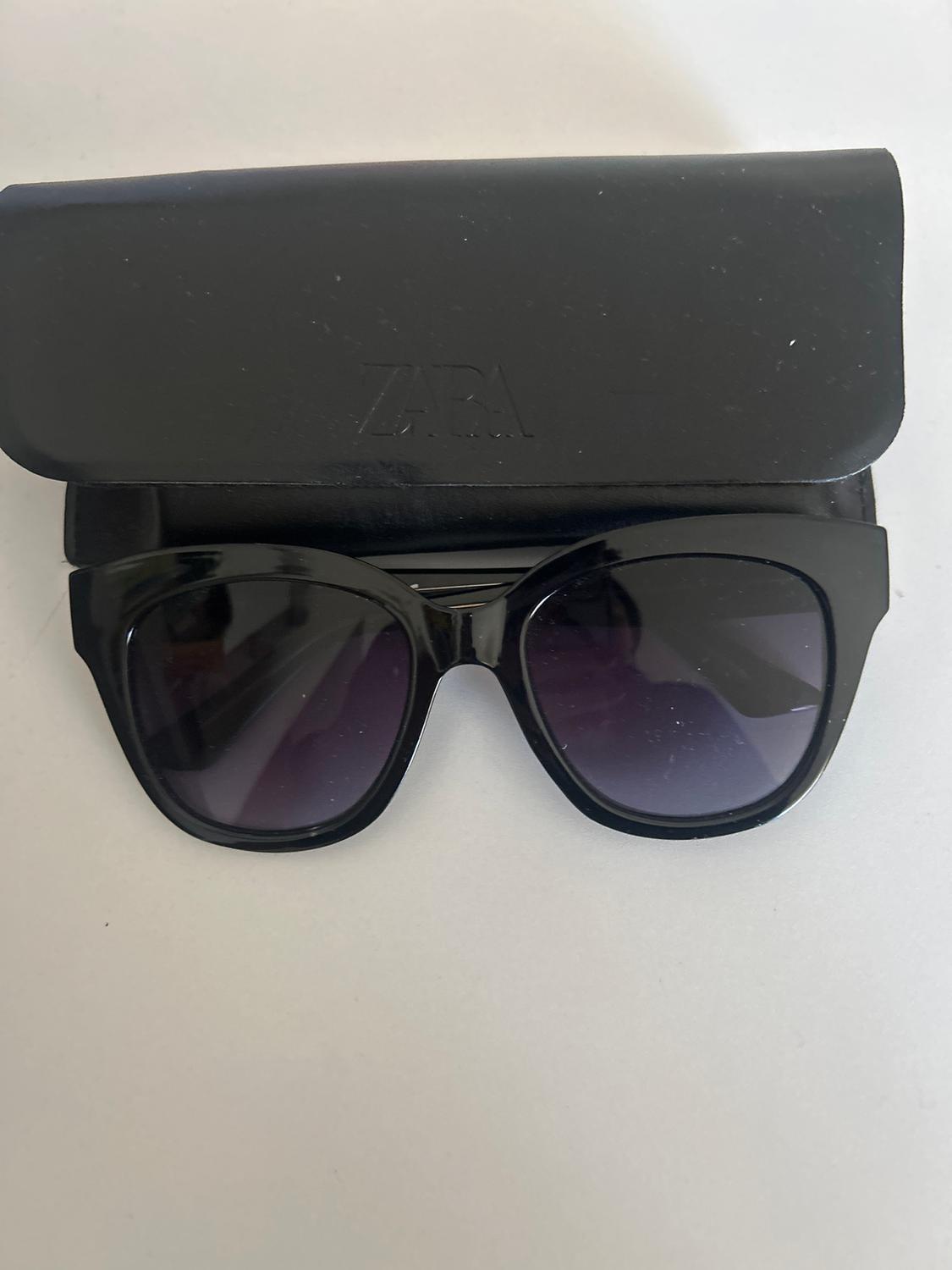 Black sunglasses with a thick frame and dark lenses, placed in front of a black case with the "ZARA" logo.