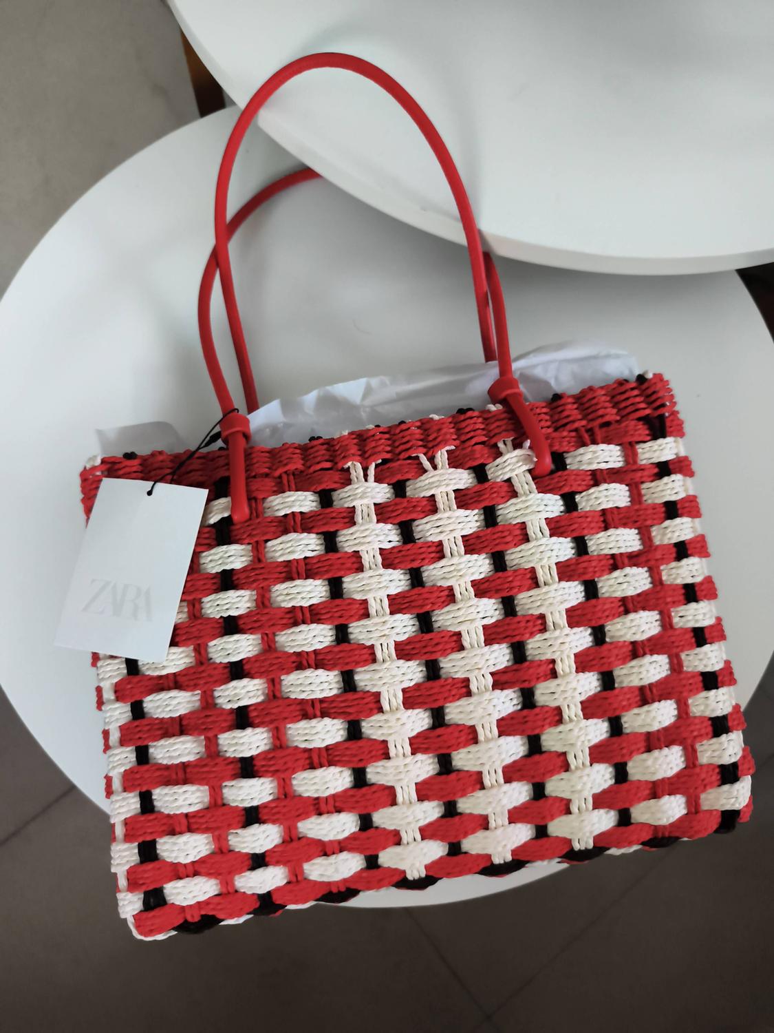 Woven bag in red, white and black with red handles on a round white table. Label with the text ZARA.
