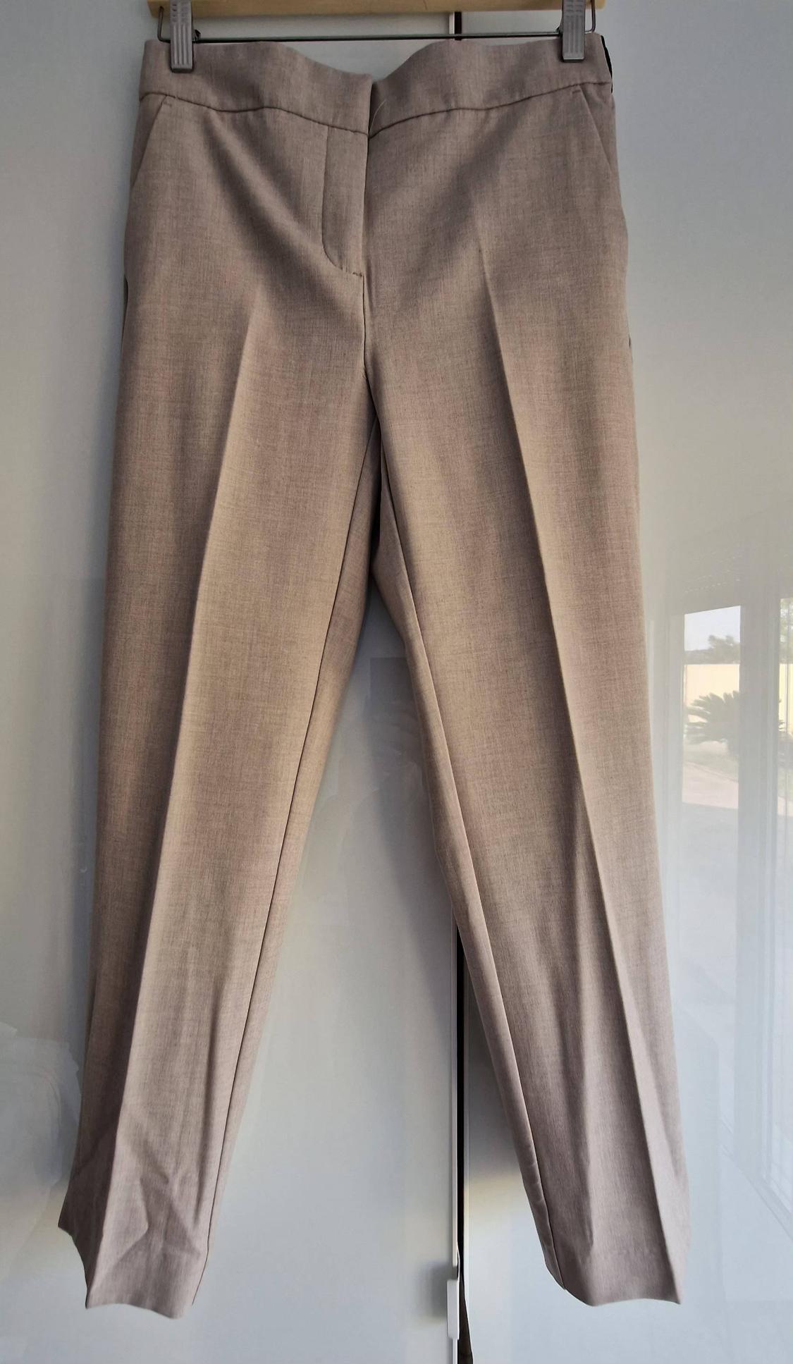 Beige smart trousers with a front crease, hanging on a coat hanger above a glossy white wardrobe reflecting a window.