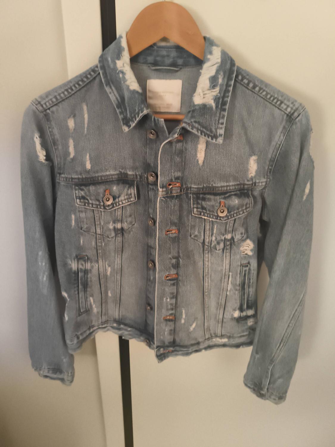 Light blue denim jacket on a wooden hanger. Its relaxed, heavily distressed style stands out, with multiple rips, frayed edges and buttons.