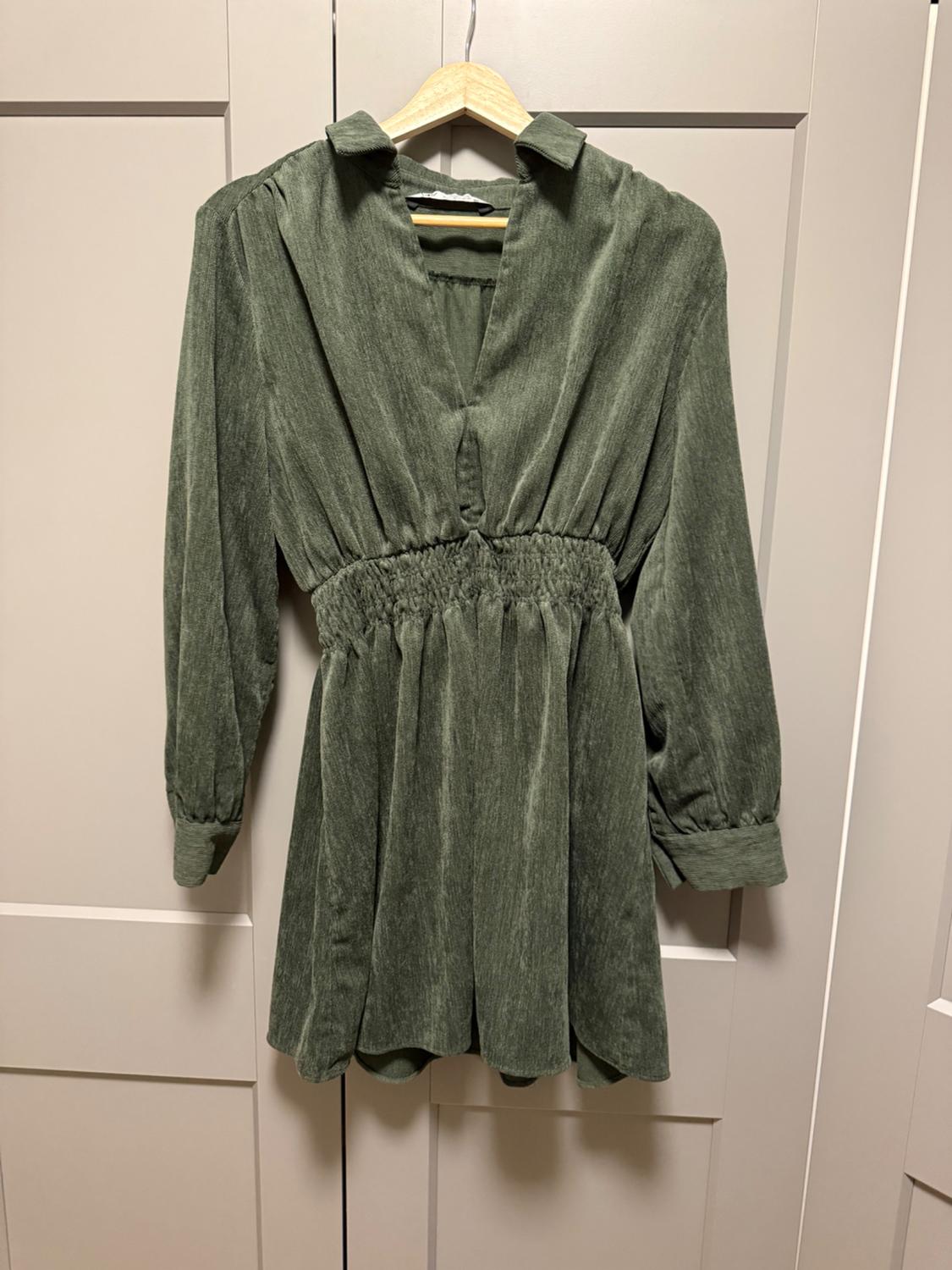 Short green long-sleeved dress with a V-neck and gathered waist, hanging on a wooden hanger in front of a light-colored wardrobe.