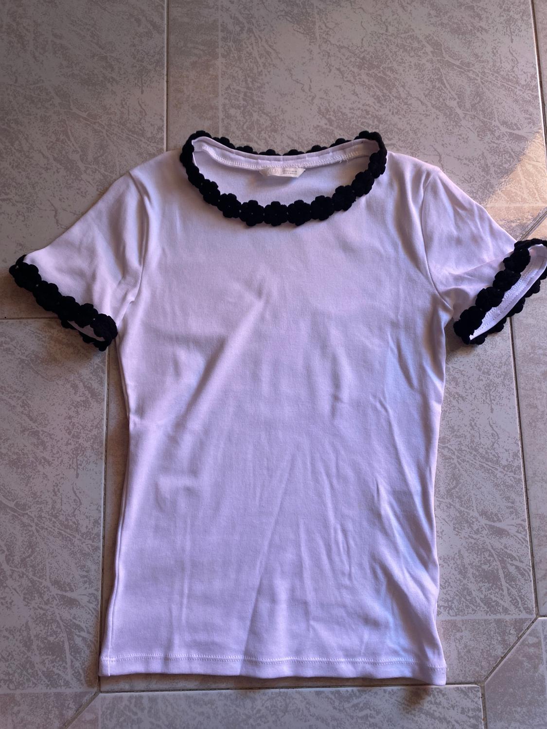 Λευκό t-shirt με μαύρες φλοράλ λεπτομέρειες στον γιακά και στα μανίκια, πάνω σε ένα γκρι πλακάτο πάτωμα με γεωμετρικό σχέδιο.