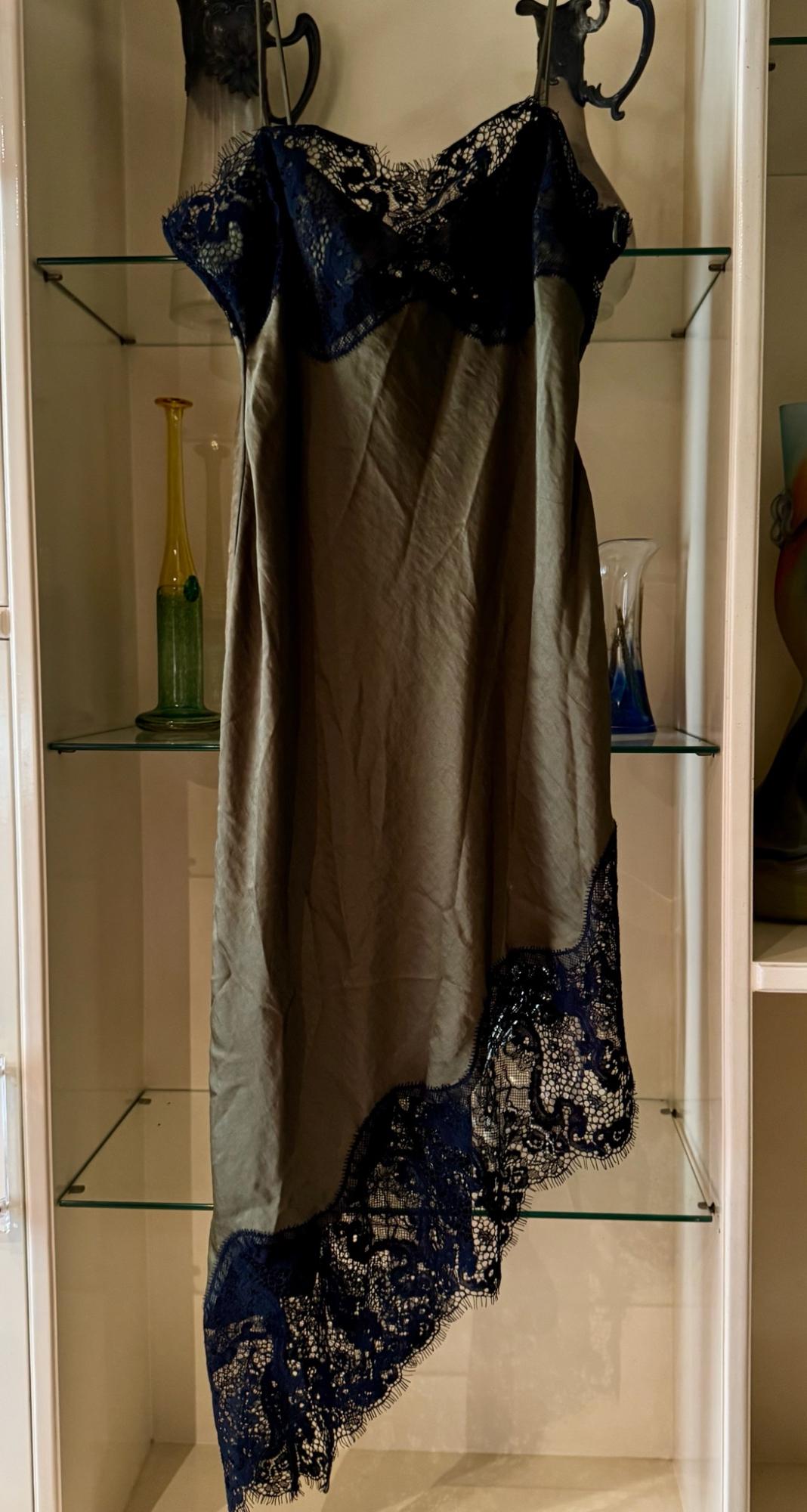 Olive-green satin slip dress with dark lace at the neckline and an asymmetric hem, hanging in front of a white display cabinet with glass shelves.
