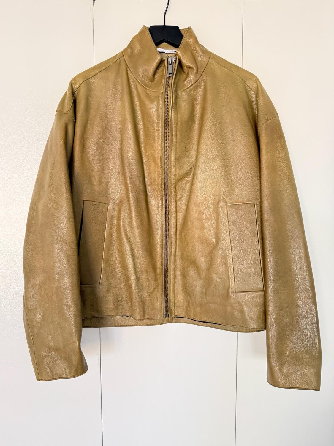 Tan jacket with a high neck, front zip and large pockets, hanging on a black hanger against a white background.