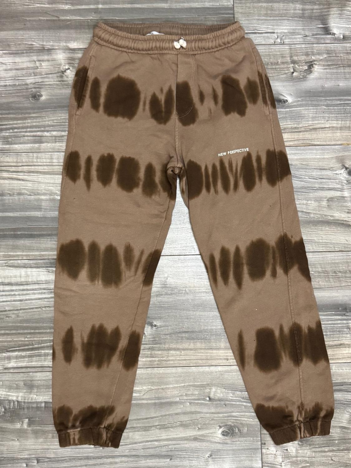 Brown jogger trousers with a dark tie-dye print, elasticated waistband and the text “NEW PERSPECTIVE” on the leg, on a wooden floor.