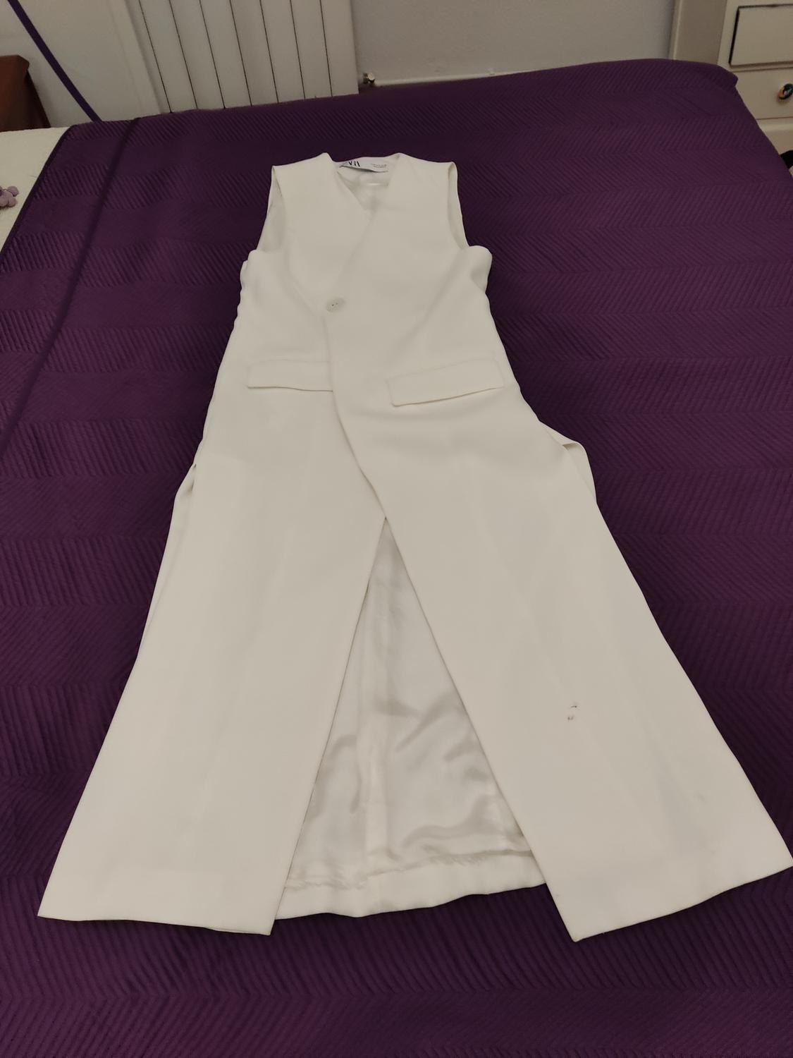 Long white sleeveless waistcoat on a bed with a purple bedspread. The waistcoat has one button and two front pockets.