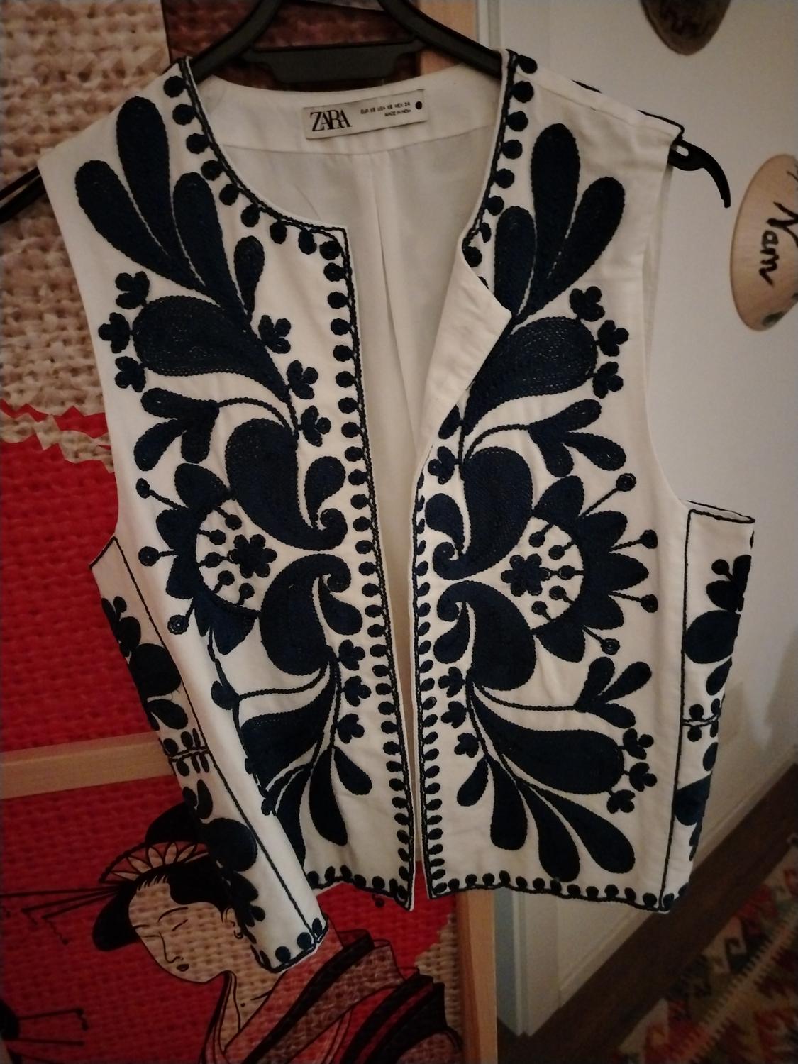 White waistcoat with black floral embroidery hanging on a hanger. Background with Japanese art and colourful rug. Visible tag: Zara.