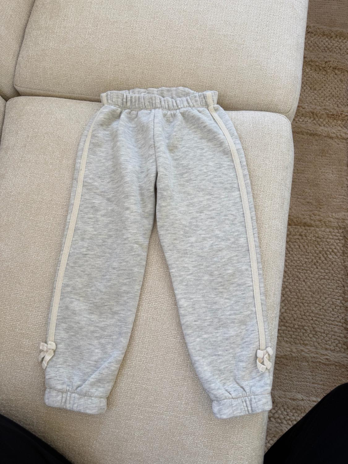 Heather gray sweatpants with an elastic waistband, side stripes, and small bows at the cuffs, on a beige sofa.