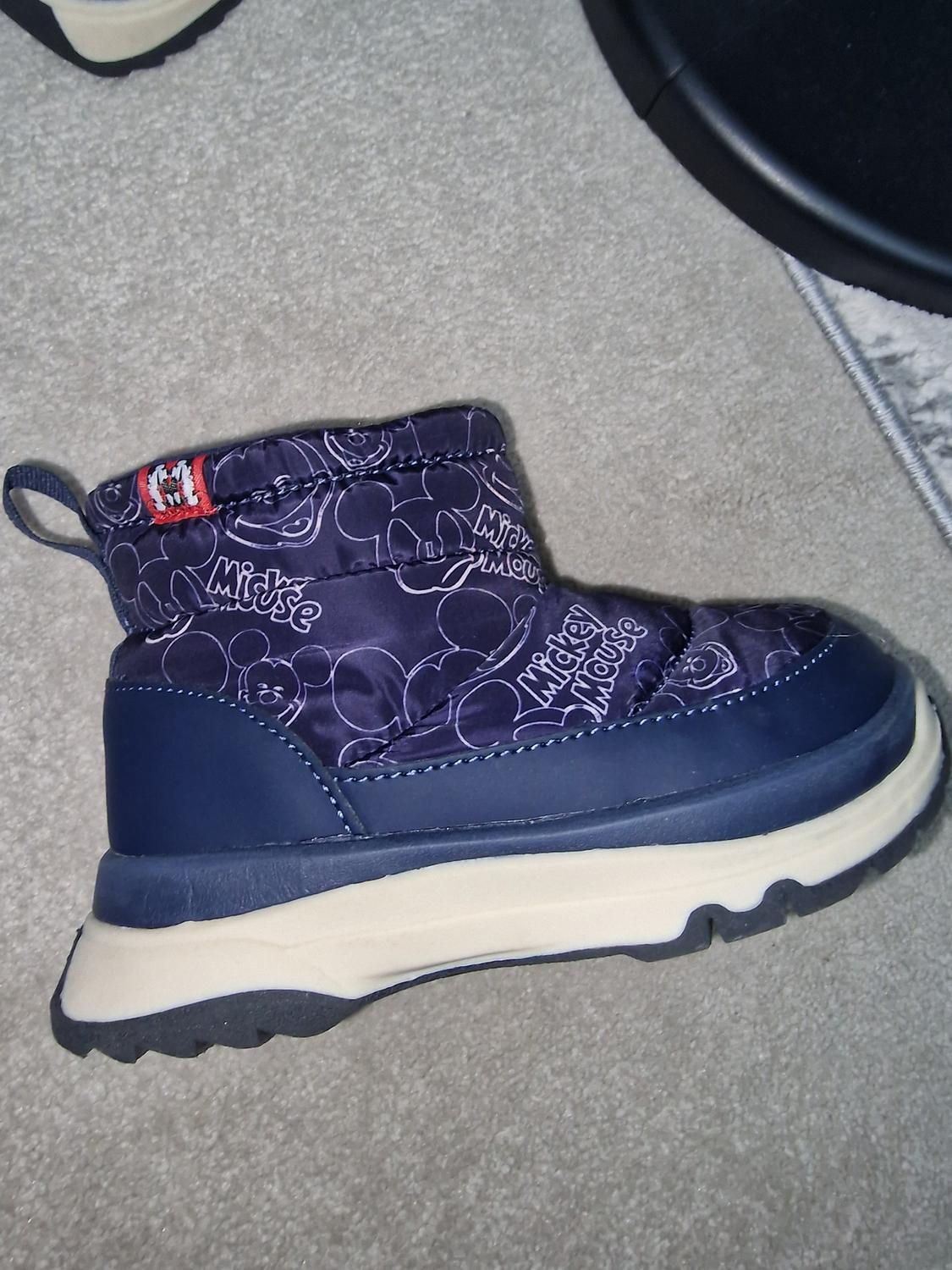 Blue child's boot with Mickey Mouse drawings on the fabric, black and white sole, on grey carpet. Visible text: "Mickey Mouse".