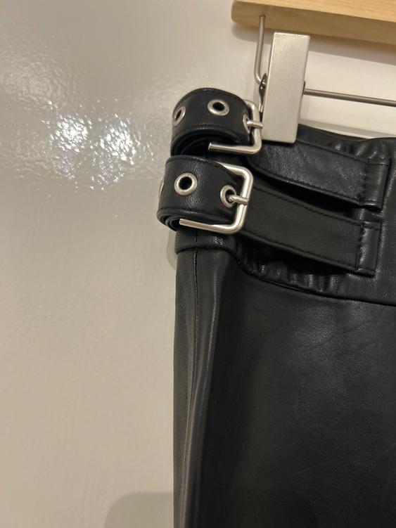 Detail of black trousers hung on a hanger, with a silver-buckled belt and metallic eyelets, on a bright white background.