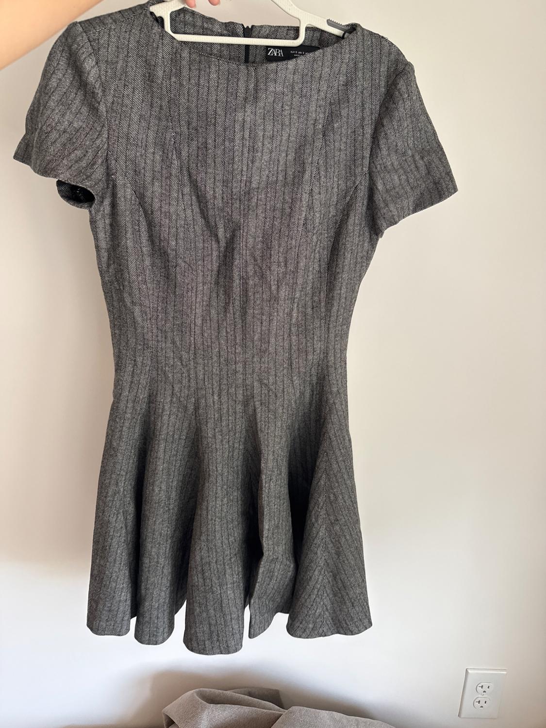 Here is a reformulated version that maintains the exact same meaning and context, without using any of the prohibited terms: "A short-sleeved gray dress with a flared skirt hangs from a hanger held by a hand in front of a white wall, leaving the ZARA label visible."