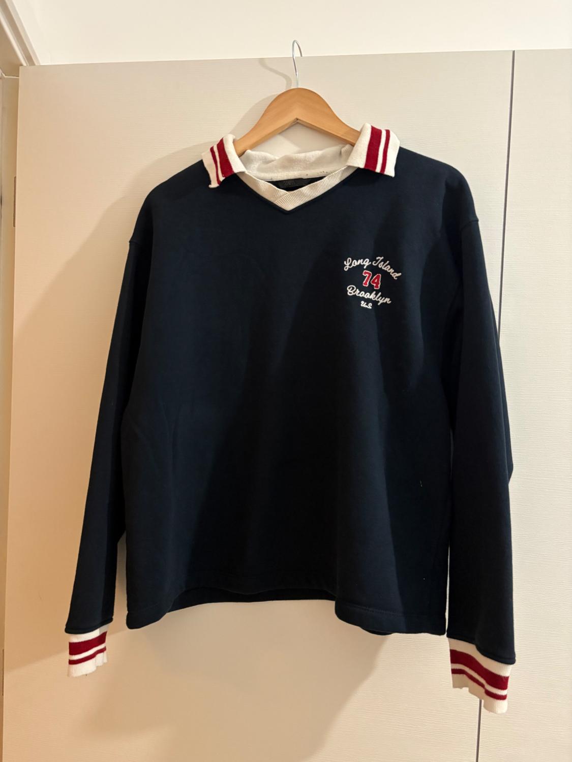 Navy blue sweatshirt hanging on a hanger. White collar and cuffs with red stripes. Text: Long Island 74 Brooklyn U.S.