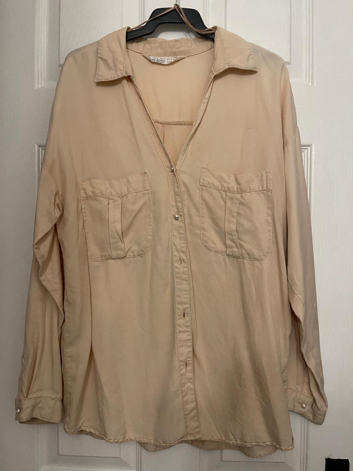 Beige long-sleeved shirt hanging on a black hanger, with two front pockets and buttons, on a white door.