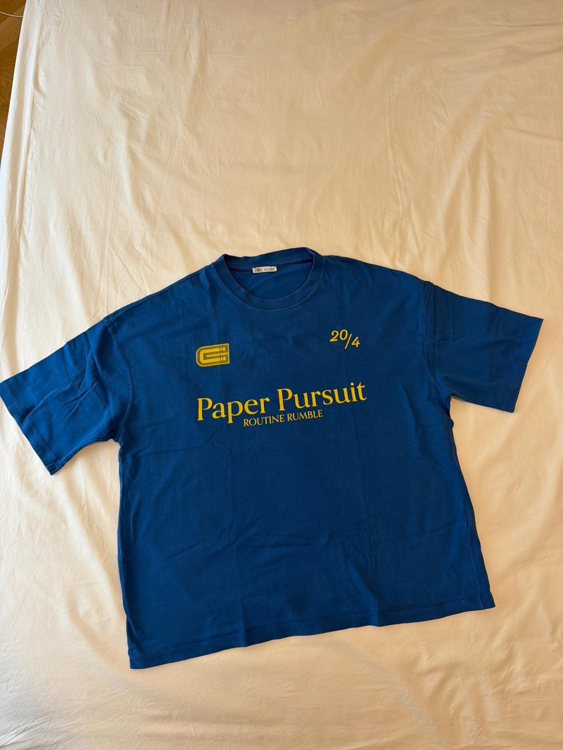 Blue short-sleeved t-shirt on a crumpled white sheet. Yellow text: 20/4, Paper Pursuit, ROUTINE RUMBLE. Relaxed style.