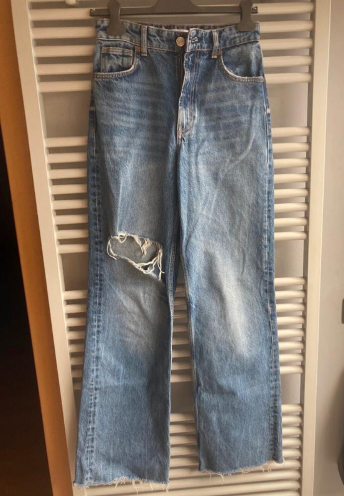 Worn blue jeans hanging on a hanger, with a rip on the left leg and frayed hem. White slatted background.