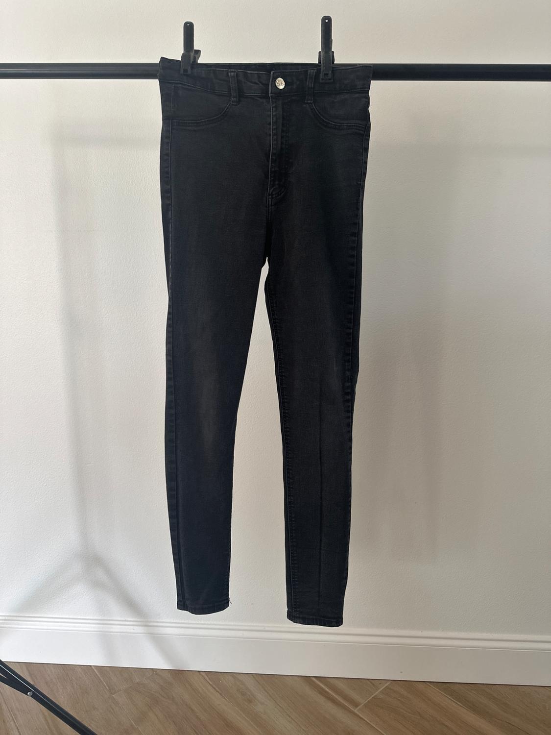 Black skinny trousers hanging on a black hanger against a white backdrop, with wooden flooring visible at the bottom.