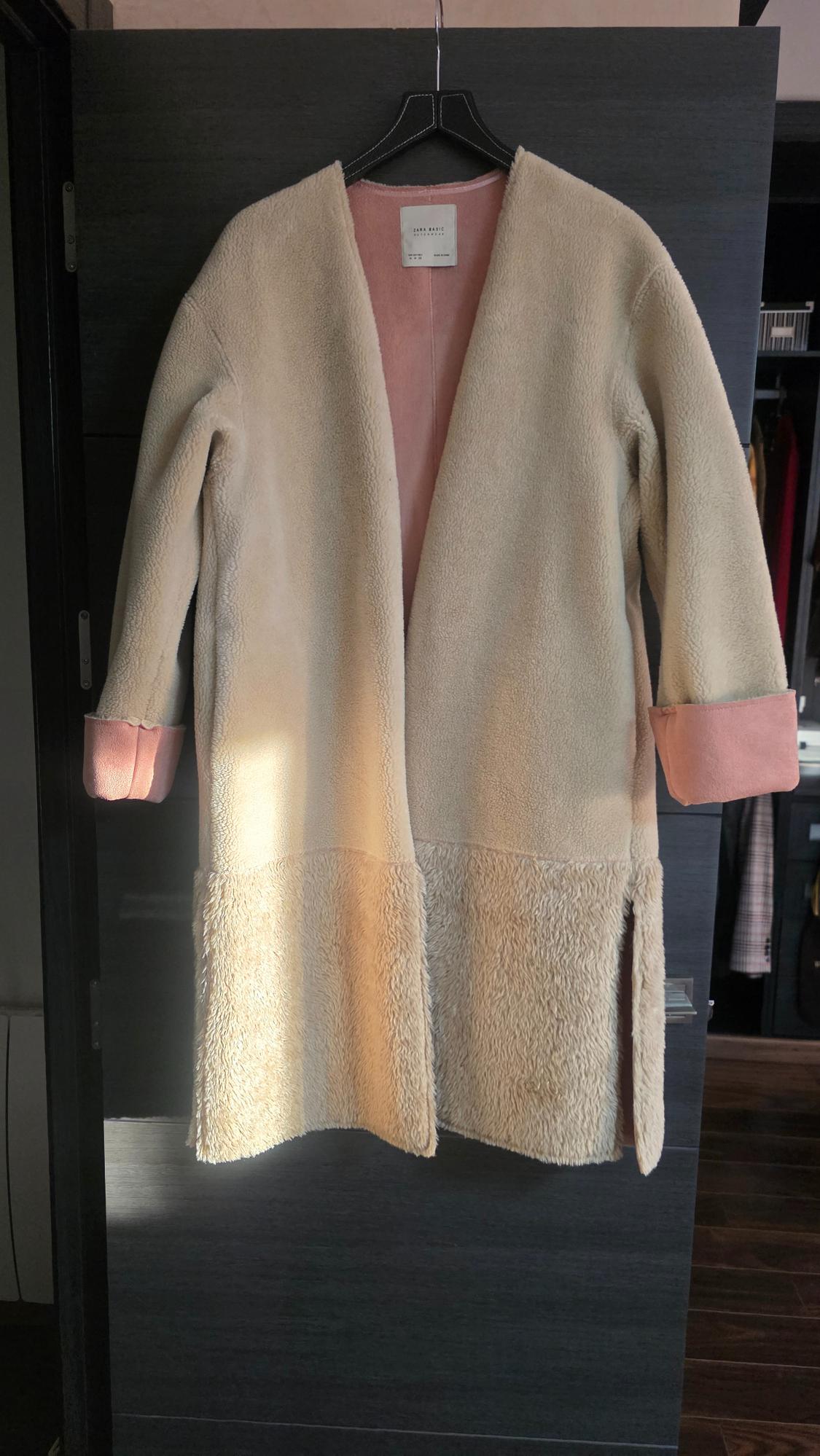 Beige, soft fabric coat with a pink interior, hanging on a black hanger. Natural light illuminates the garment from the left.