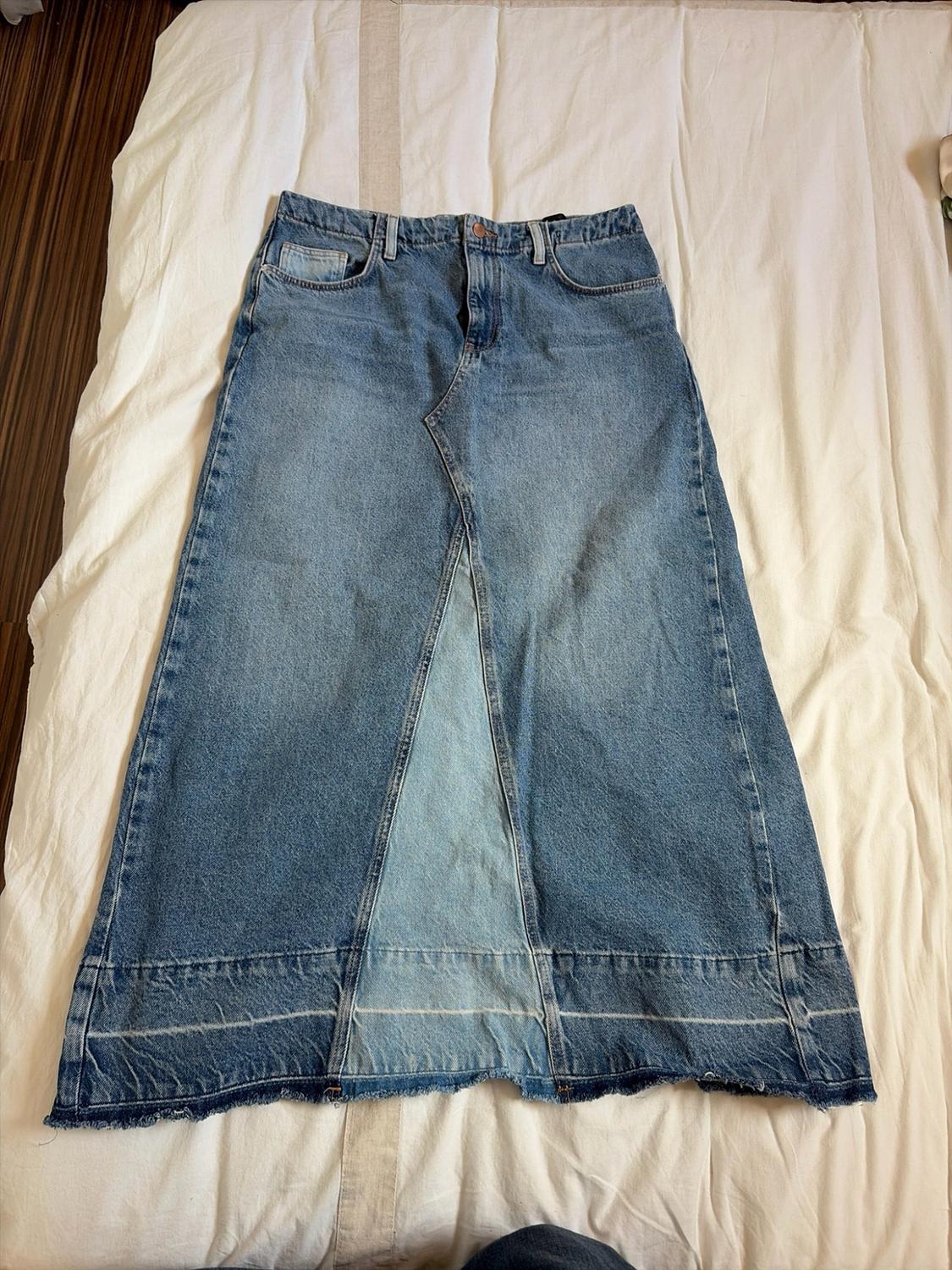 Blue denim maxi skirt with a distressed wash, lighter front panel and frayed hem, laid out on a white sheet.