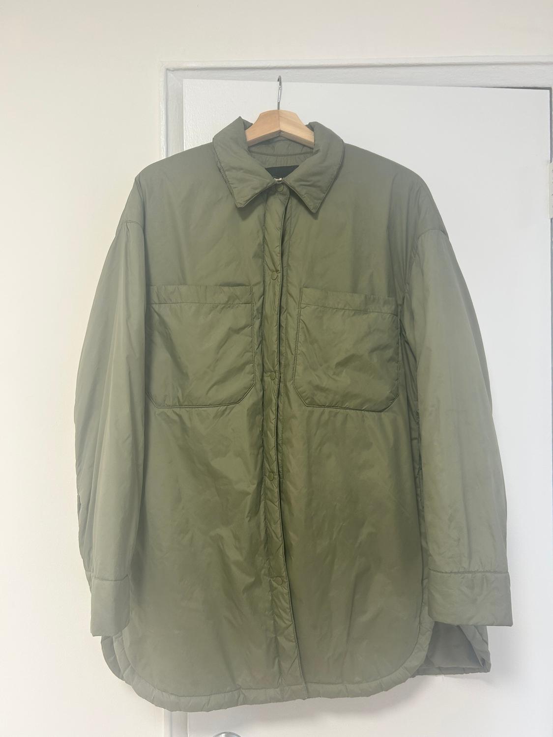 Olive green quilted jacket hanging on a wooden hanger over a white door. Simple design with front pockets.