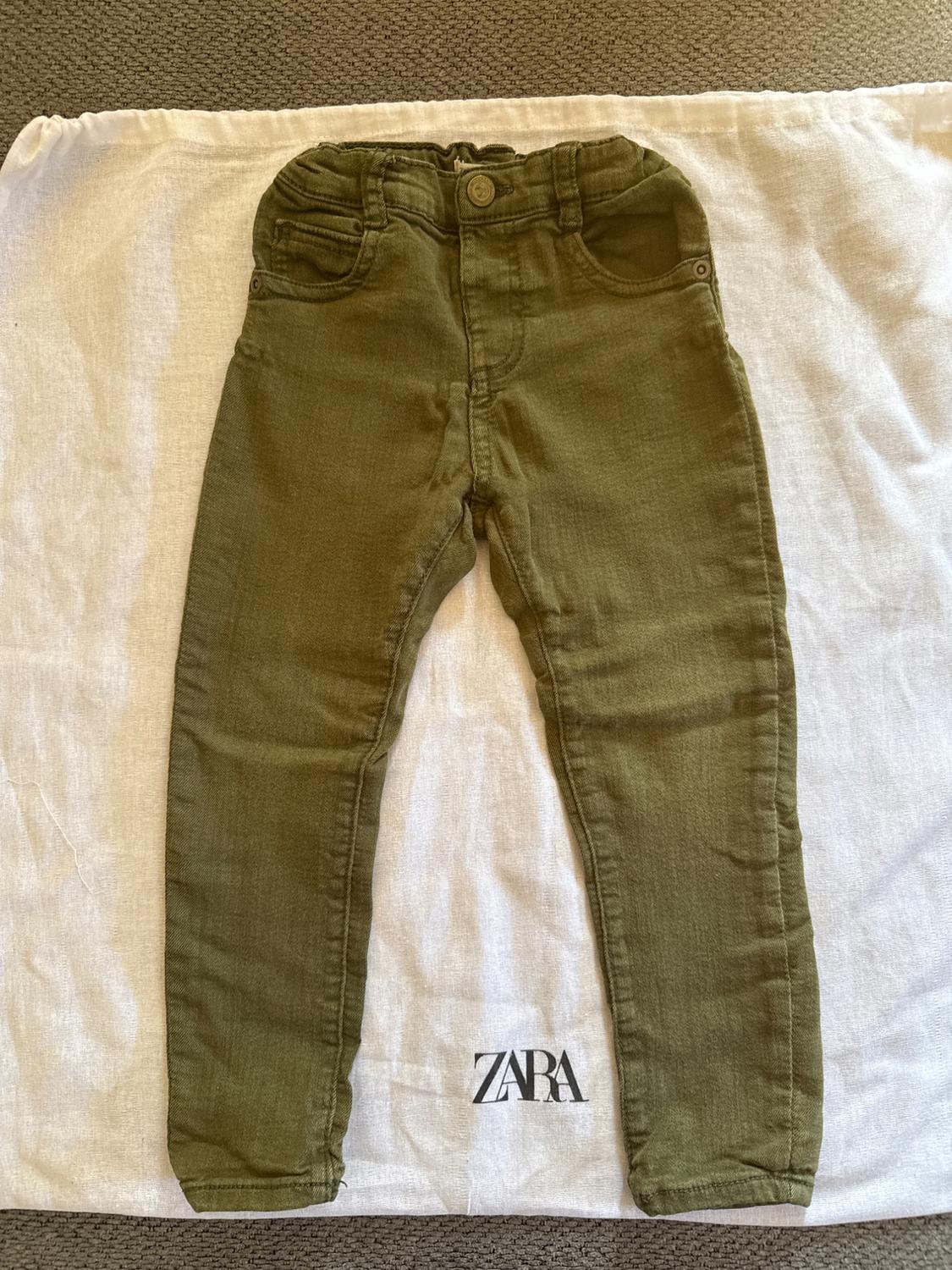 Green pants for children on white fabric with "ZARA" logo visible at the bottom. Gray carpet background.