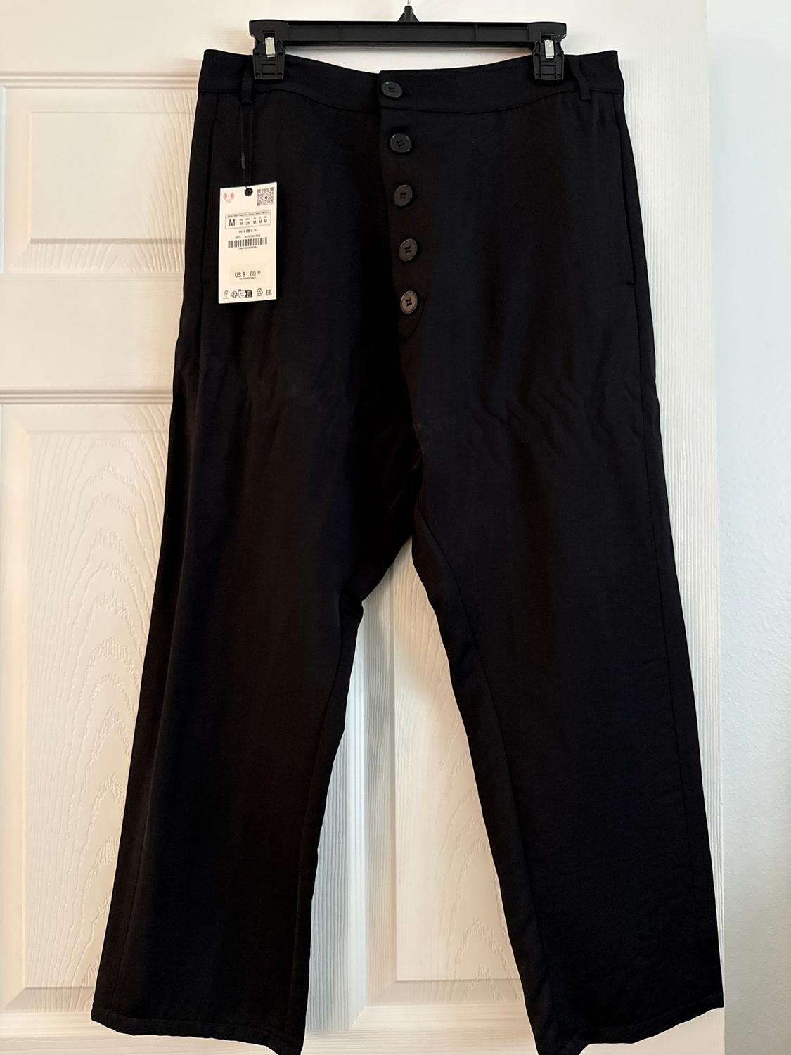 Black trousers hanging on a hanger with front buttons and a visible tag. White door background.